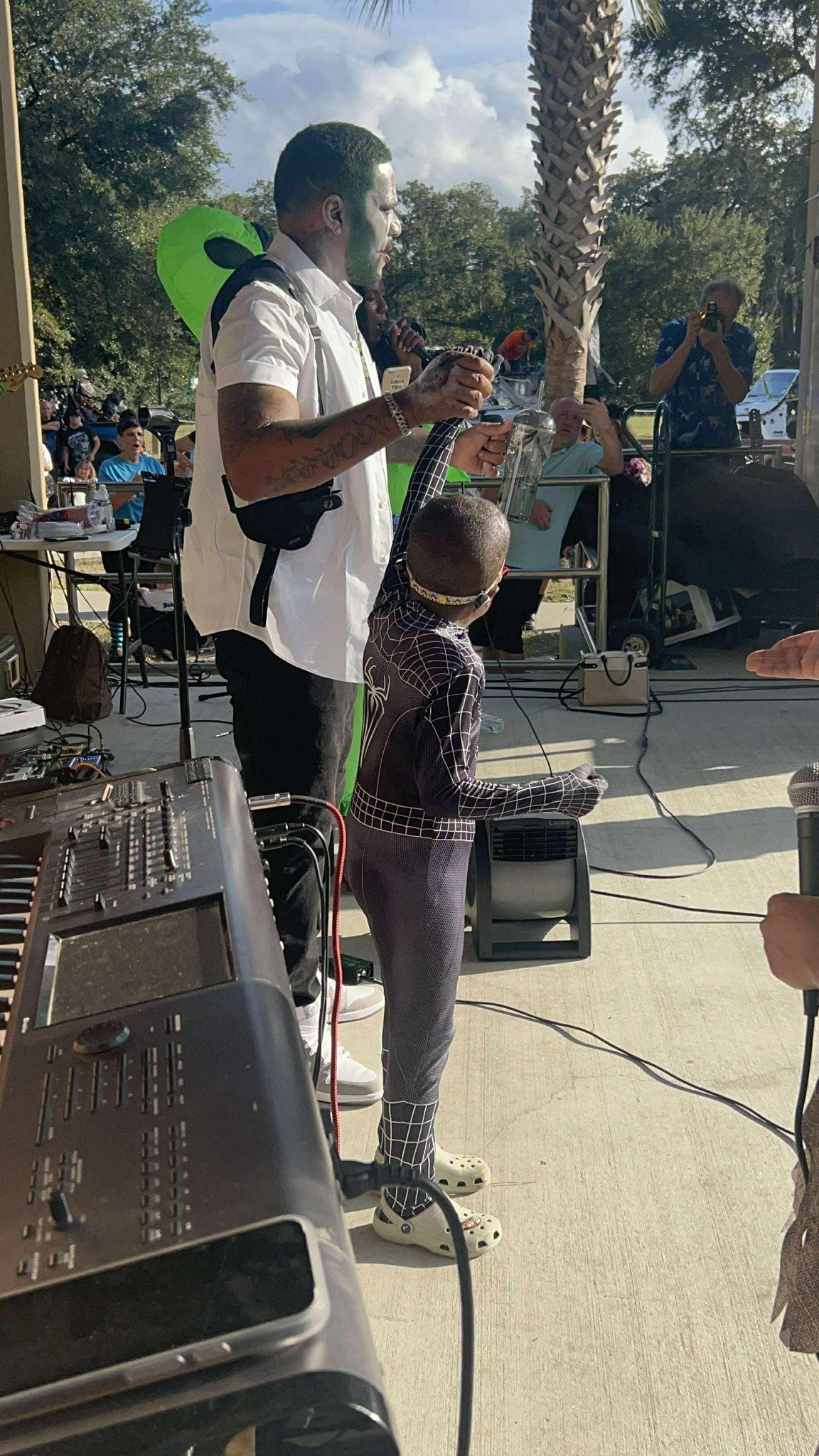 A man with painted face in a white shirt holding a microphone at an outdoor event, with a young boy dressed in a Spider-Man suit standing nearby. There are people seated in the background, some taking photos, along with DJ equipment and a palm tree.