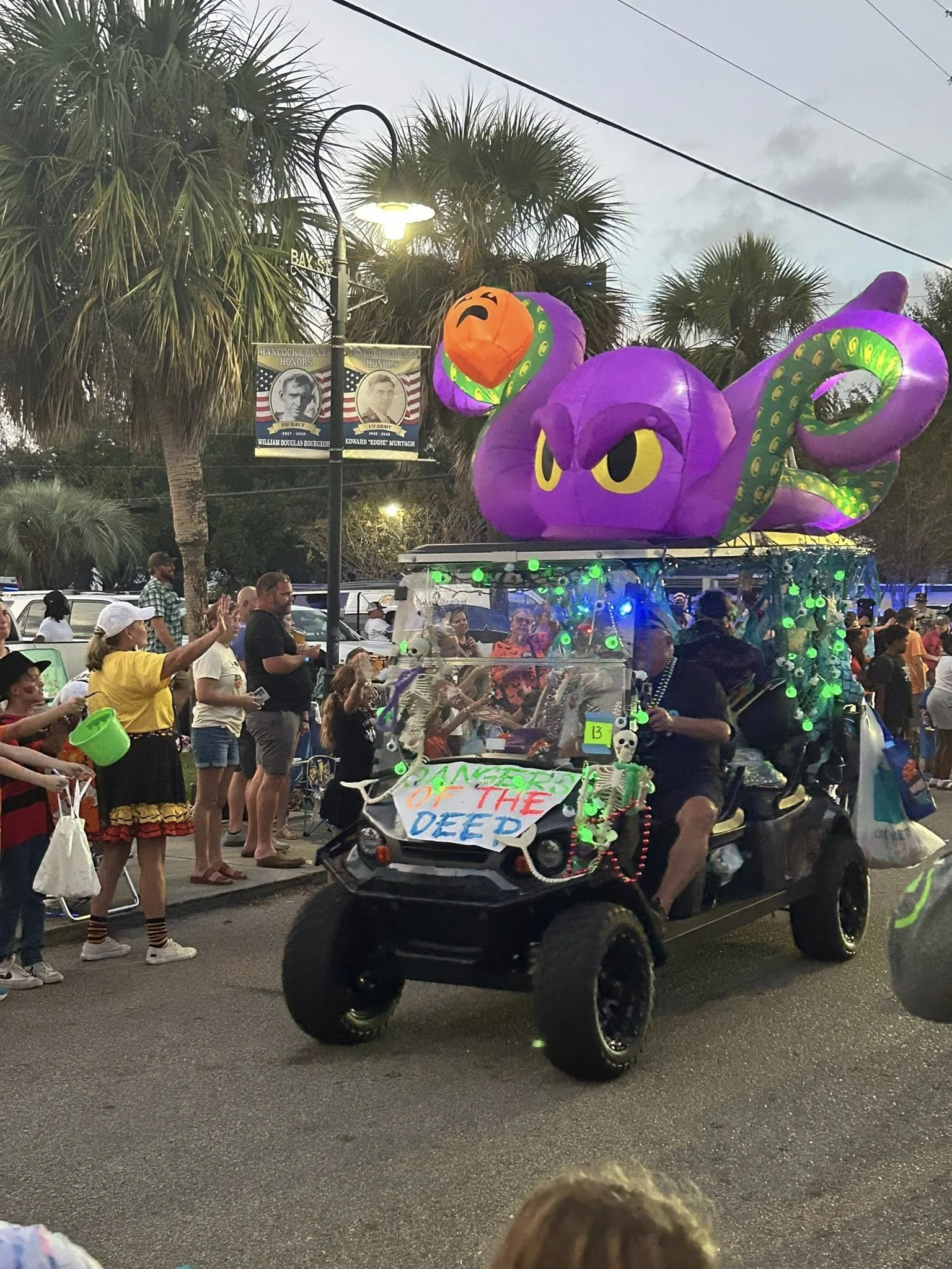 Parade float decorated with a purple octopus and a purple cartoon octopus with yellow eyes, surrounded by people and palm trees.
