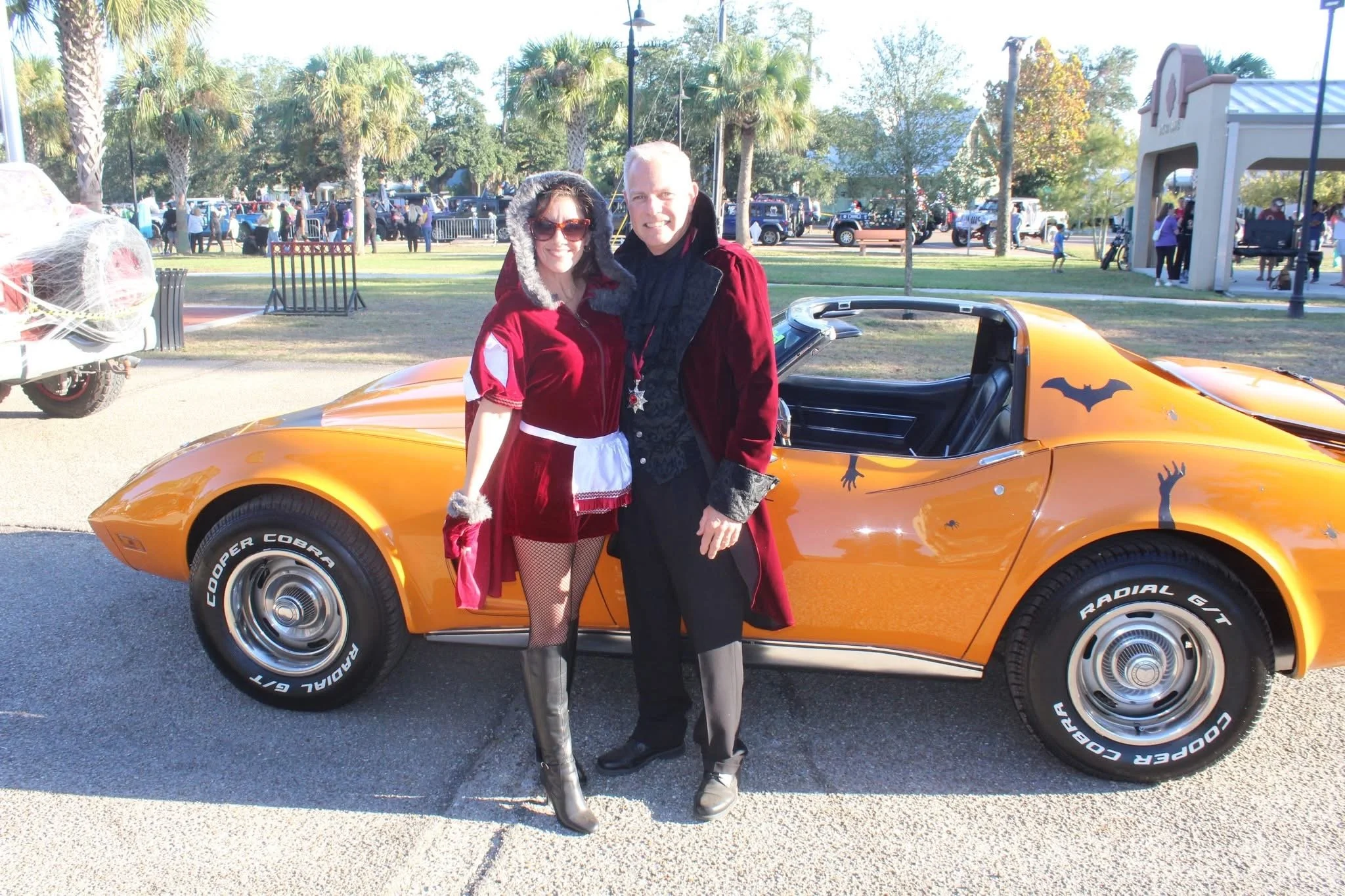 A woman in a red cloak and fishnet stockings standing next to a man dressed as a vampire, both smiling, in front of a yellow sports car with Halloween decorations at an outdoor event with trees, people, and cars in the background.