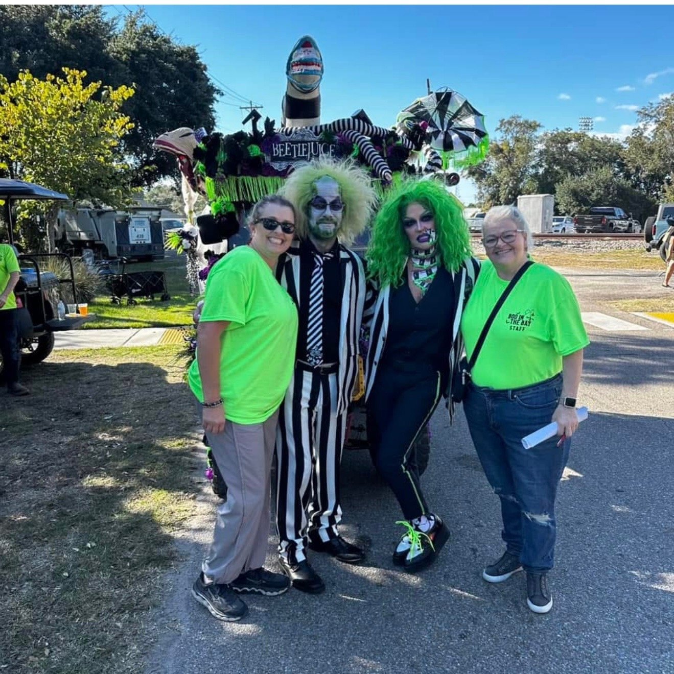 Four people standing outdoors, three women and one man. Two women and one man are dressed in black and white striped costumes, with colorful makeup and wigs. One woman is wearing a bright green wig and costume. The third woman is wearing a lime green
