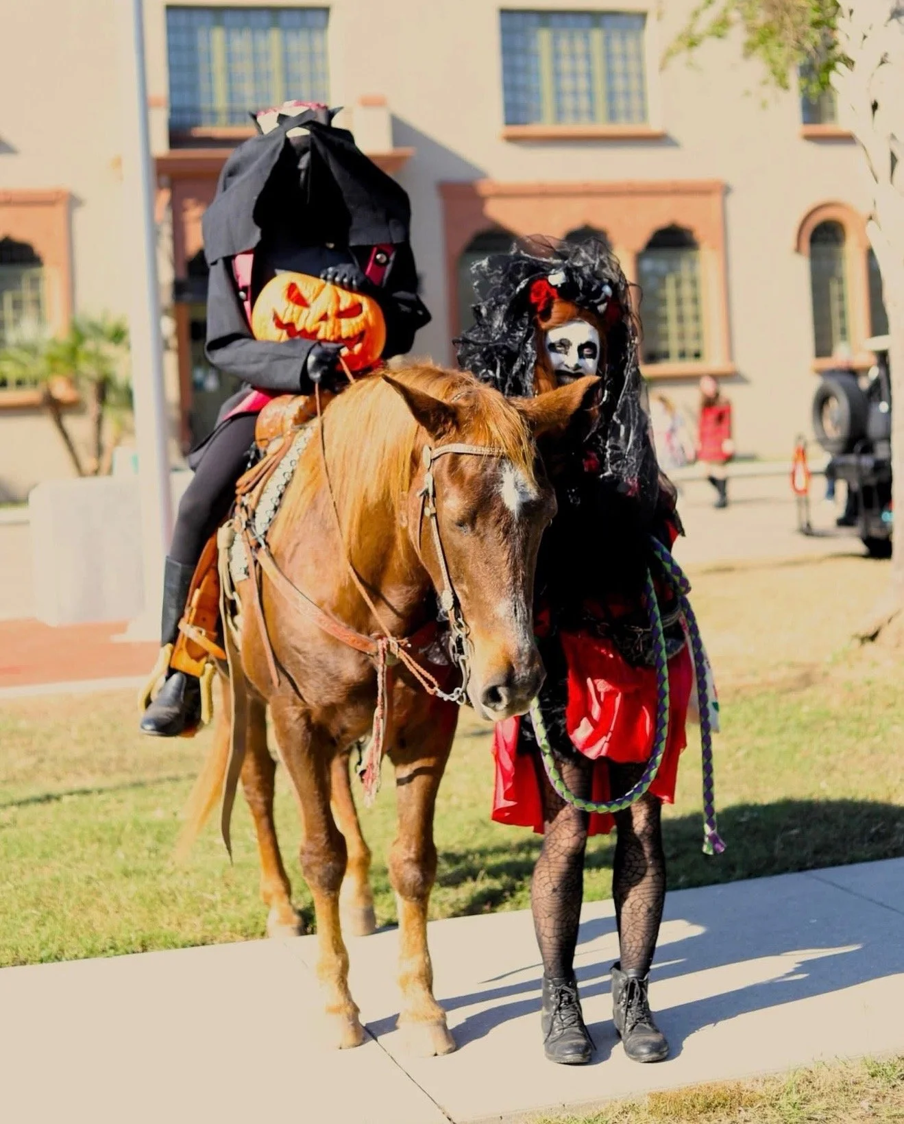 People in Halloween costumes, including a person dressed as a witch riding a horse and another dressed as a spooky character with face paint and black lace headdress, standing outside near a building.