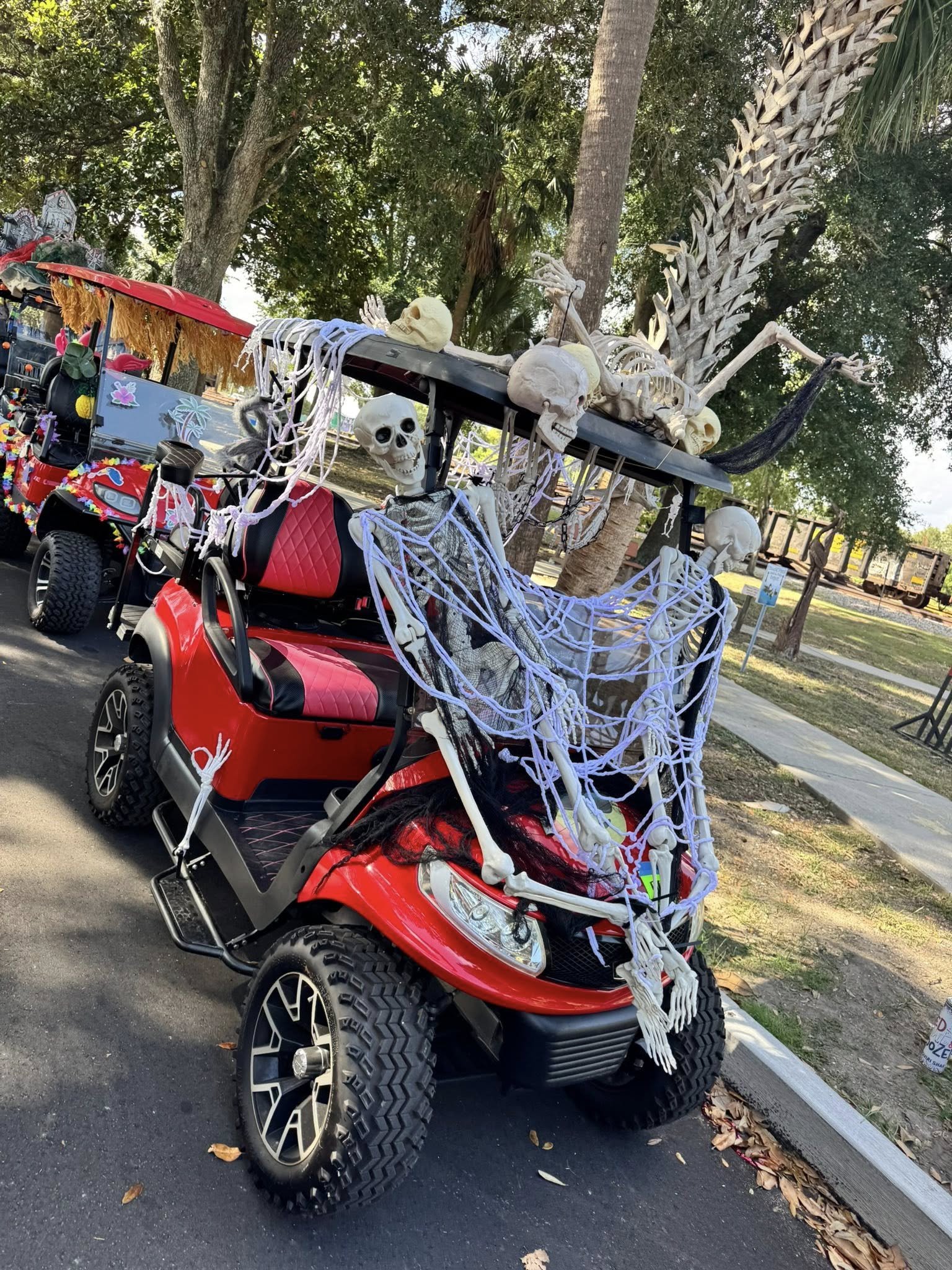 Golf cart decorated with skeletons, skulls, bones, skull head, white tendrils, and tropical flower designs, parked on a paved road near trees.