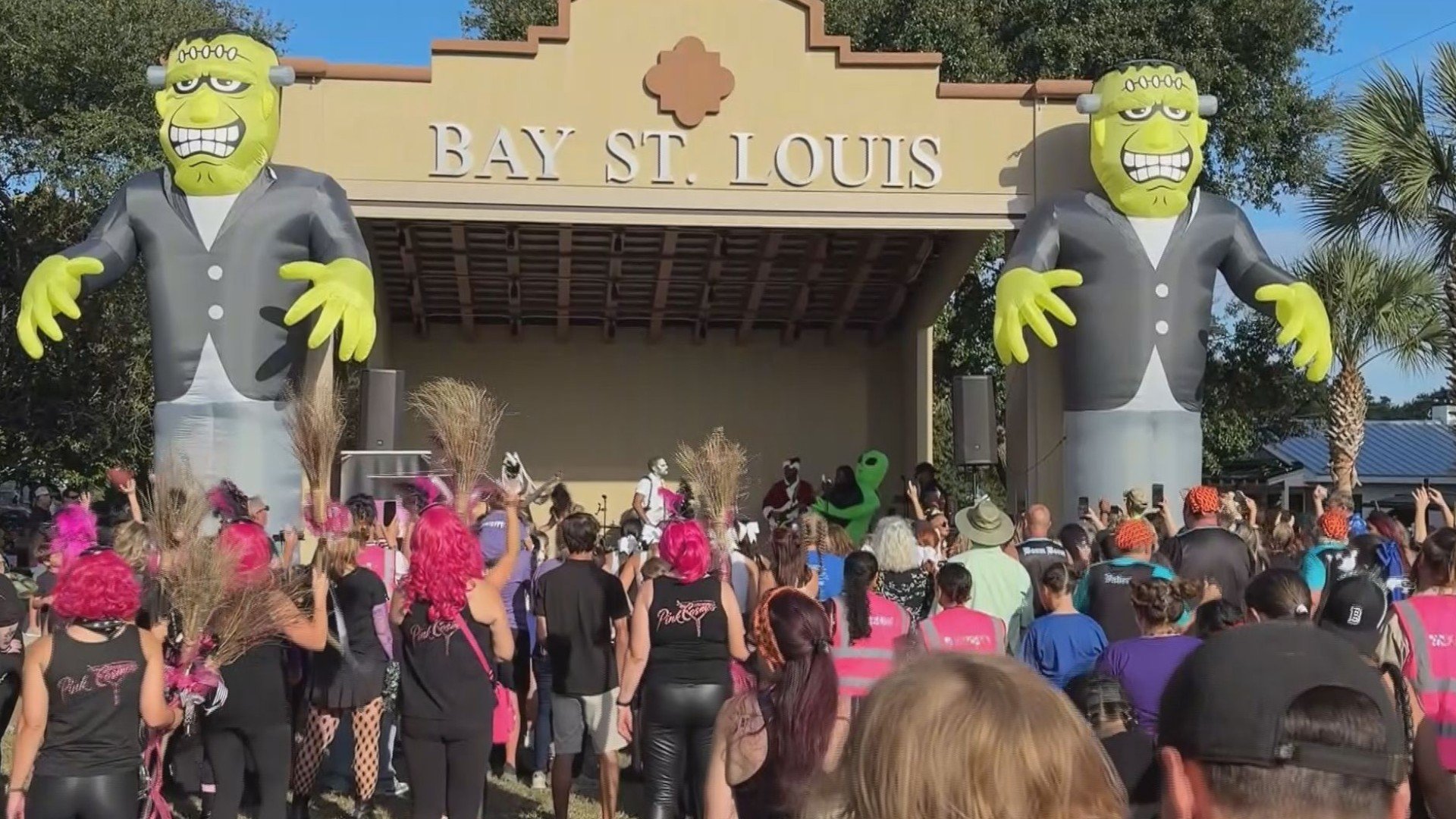 Concert stage at Bay St. Louis with two large inflatable figures resembling Frankenstein. Audience has people with pink hair and costumes, some in black shirts with pink logos, gathered in front of the stage in a sunny outdoor setting with palm trees