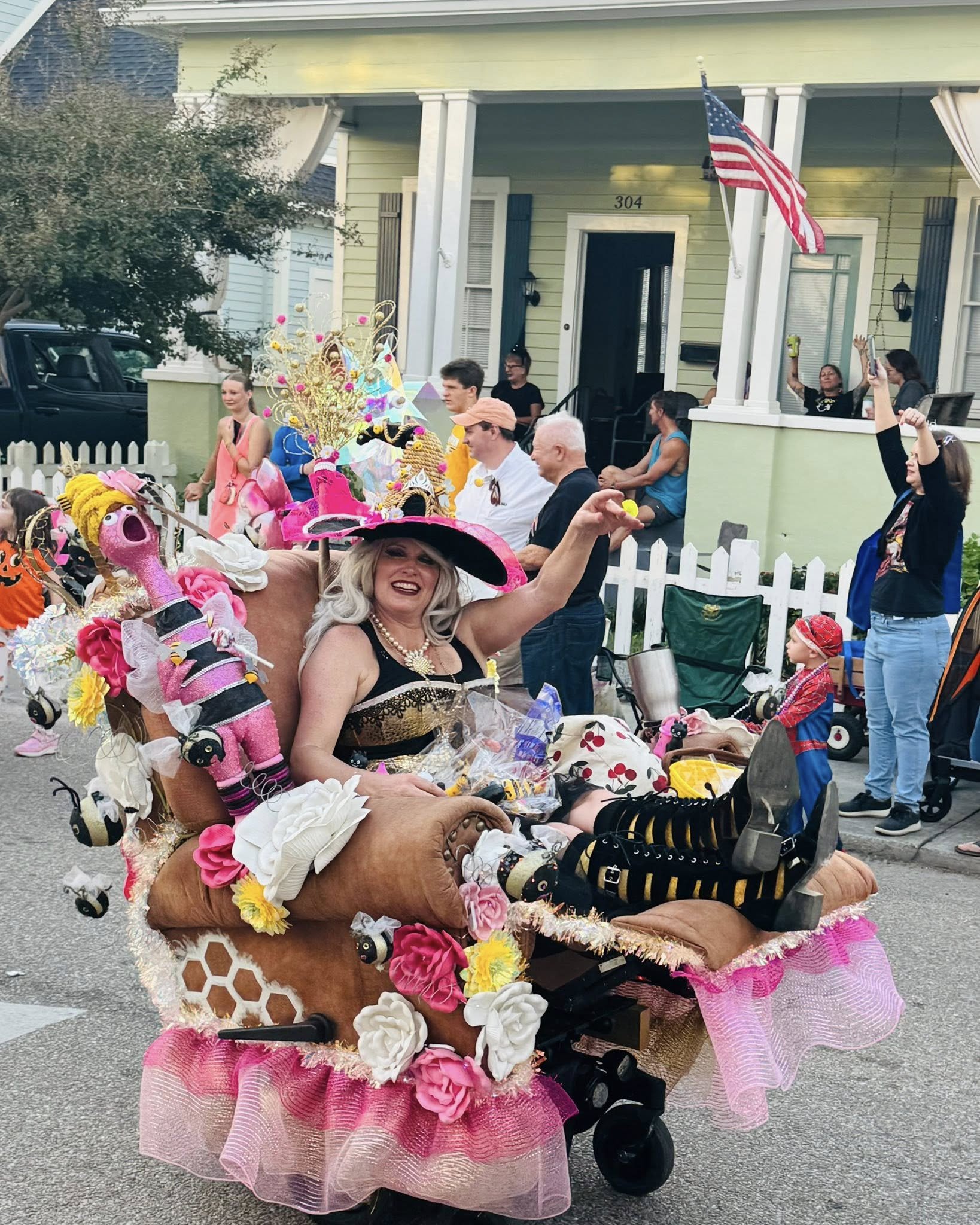 A float decorated with pink, yellow, and white flowers, pink and black stuffed animals, and colorful ribbons, being pushed down a street in a parade. People are seated or standing nearby, and a house with a front porch and an American flag is visible