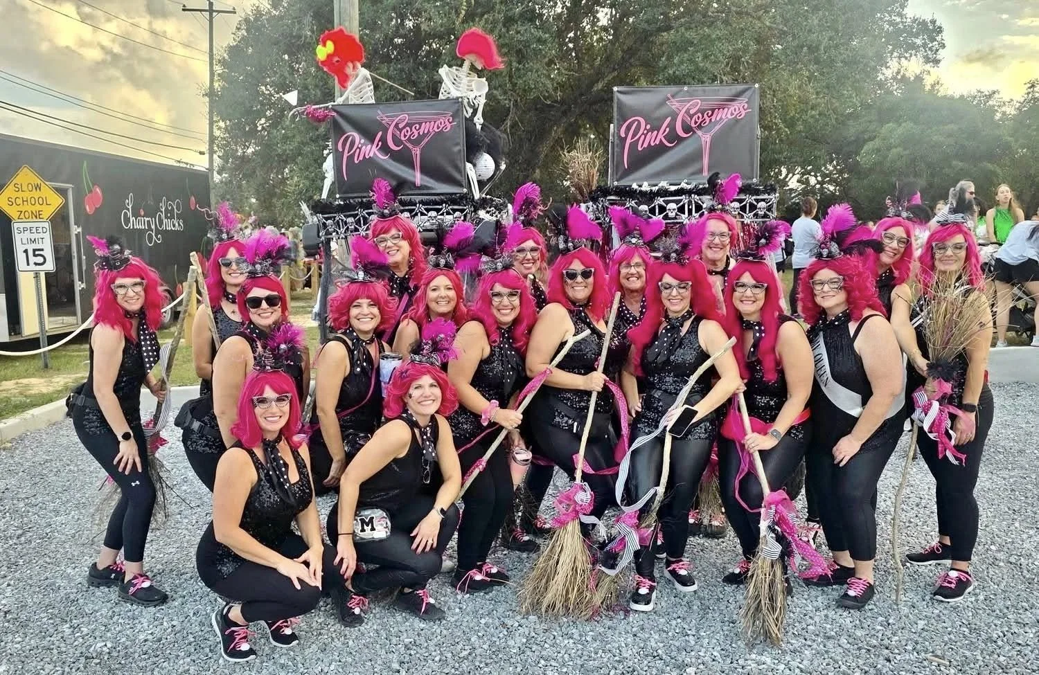 A group of women dressed as witches with bright pink hair and costumes, standing in front of a parade float decorated with skeletons and signs reading "Pink Cosmos." They are outdoors on a gravel surface, smiling and holding broomsticks and other Hal