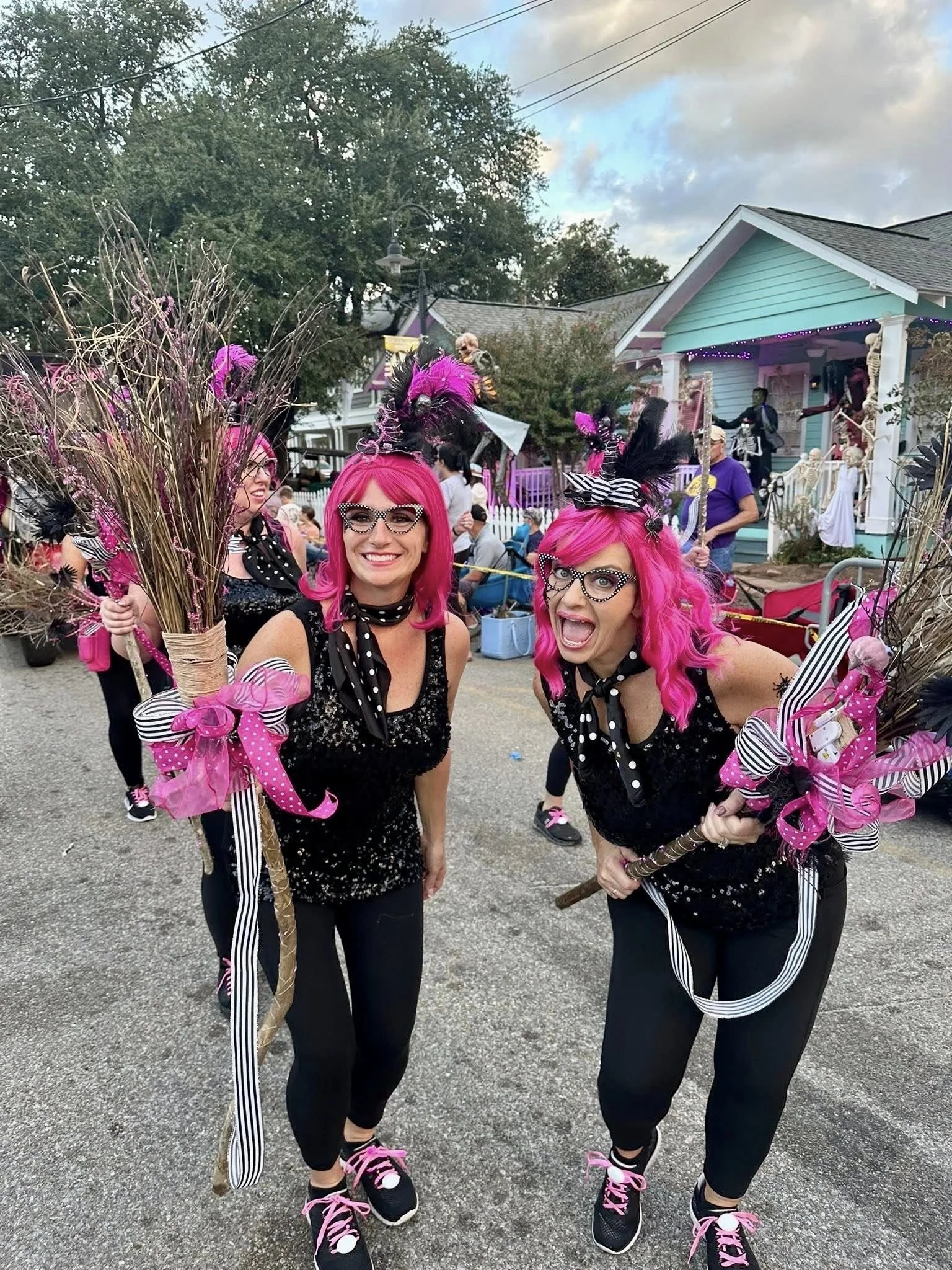 Two women with pink hair smiling and holding witch brooms at a Halloween event. They are wearing black outfits with polka-dot scarves and pink laces on their shoes, along with glasses and costumes with feathers and bows. In the background, there are 