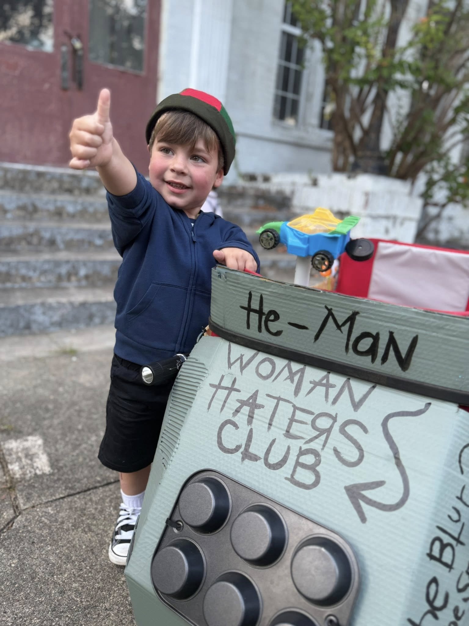 A young boy wearing a black hat with red and green stripes, a navy blue jacket, and black shorts giving a thumbs-up. He is standing next to a decorated cardboard box resembling a speaker with handwritten words, in front of a house with steps and tree