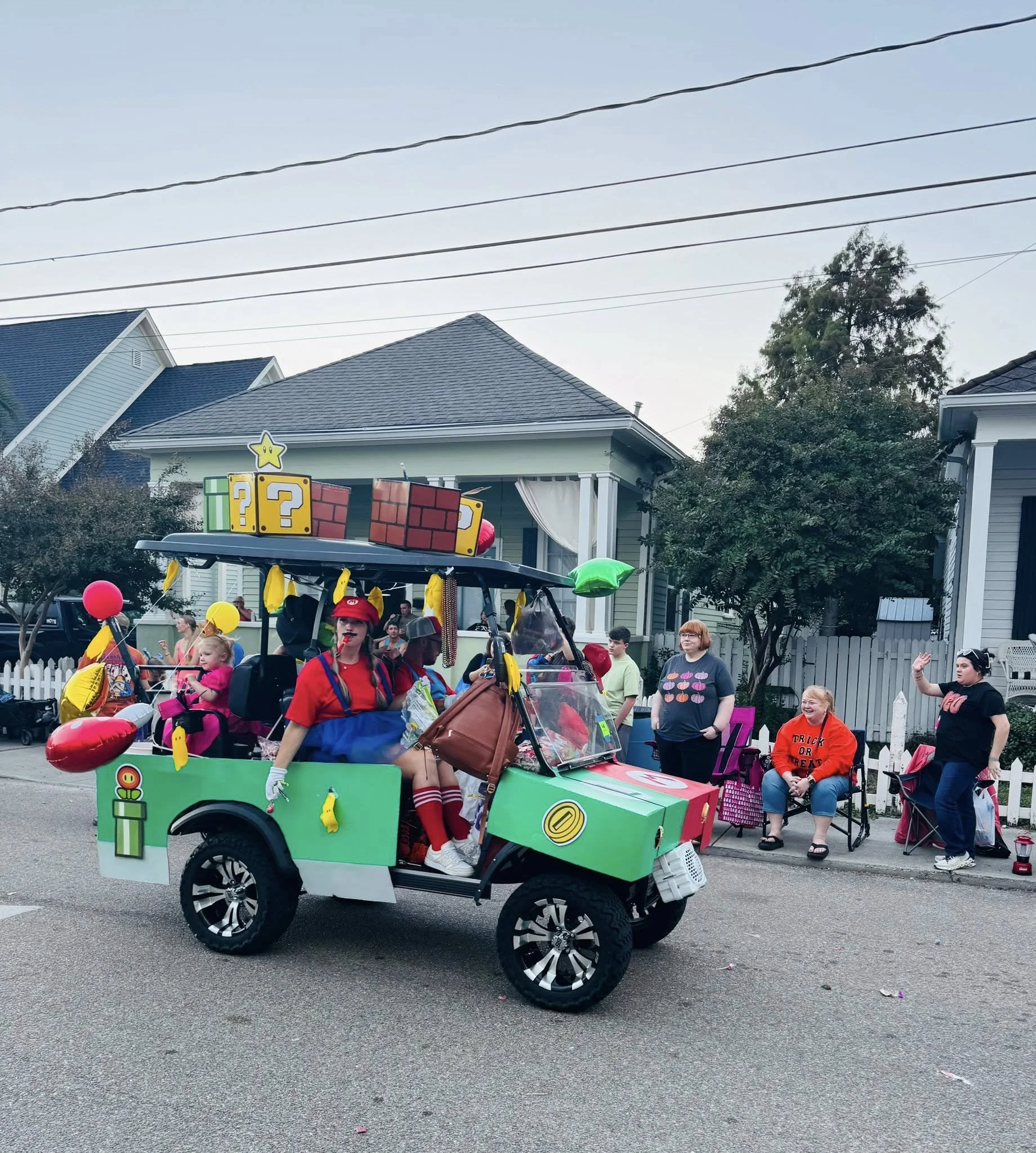 A decorated vehicle in a parade with people dressed in costumes, including a person dressed as Mario, and a float with question mark blocks from a video game. The vehicle is green with oversized wheels.