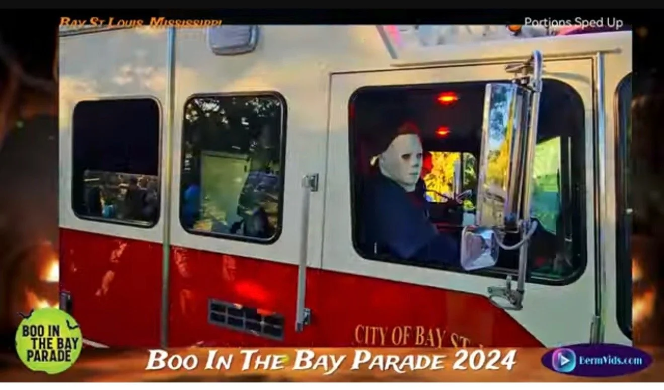 A person wearing a Michael Myers Halloween mask is driving a fire truck in a parade, with a sign indicating the event as 'Boo In The Bay Parade 2024' in Bay St. Louis, Mississippi.