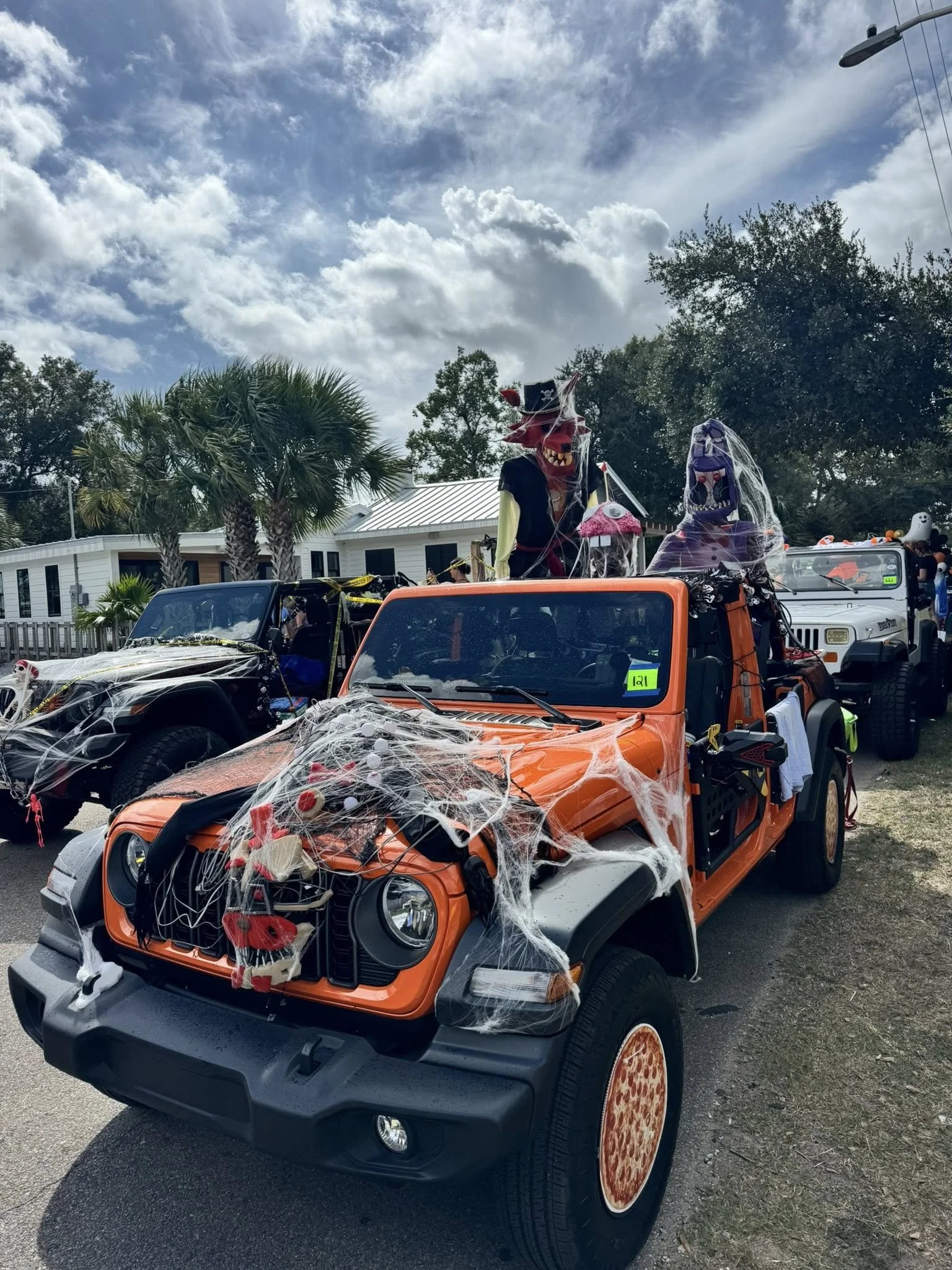 A decorated orange off-road vehicle with spider and web decorations, featuring giant pizza slice wheel covers, parked outdoors under a cloudy sky with palm trees and holiday decorations visible.