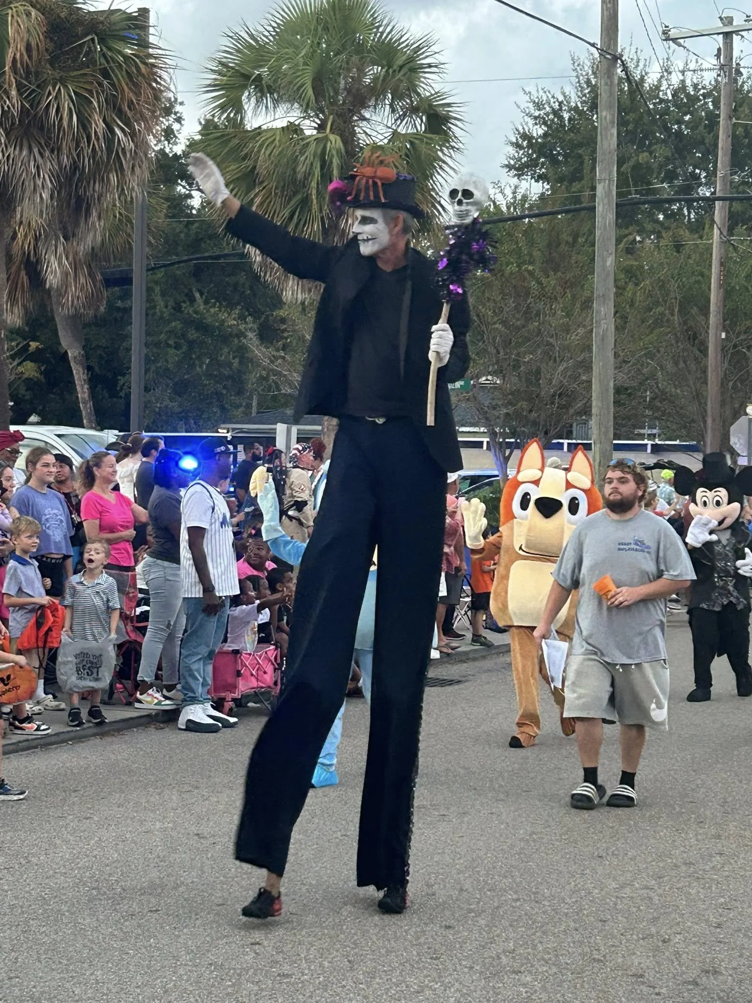 Person dressed as a skeleton in black costume with a large hat decorated with a crab and other embellishments, holding a staff with a skull, walking among a crowd at a parade or street event, with people and costumed characters including a dog mascot