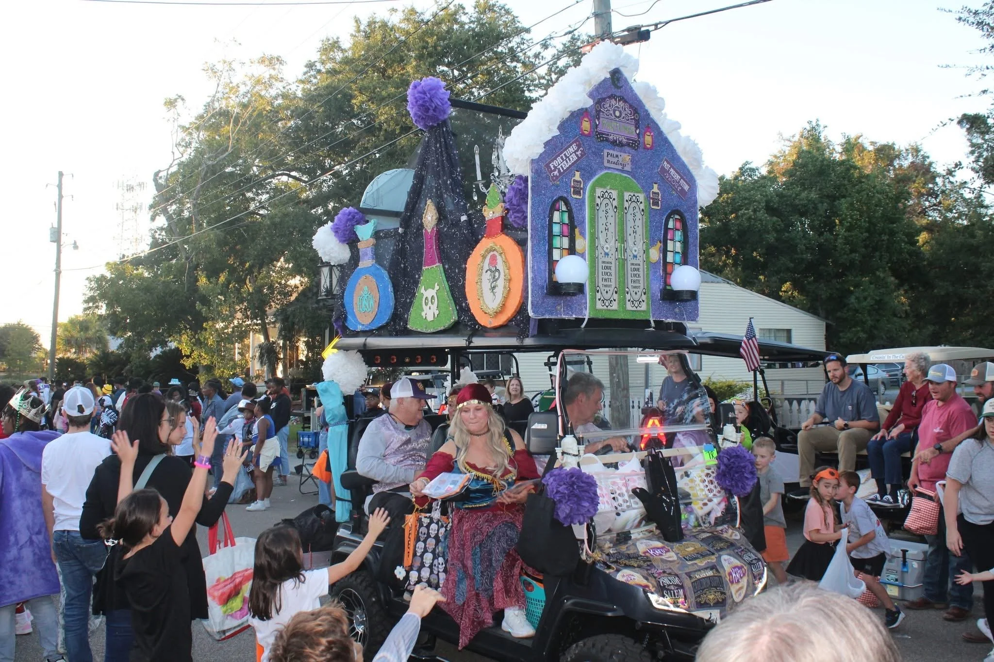 A parade float with a spooky haunted house design, decorated with purple, white, and orange accents, carrying a vendor selling merchandise. The float is surrounded by a crowd of people, including children and adults, participating in the parade on a 