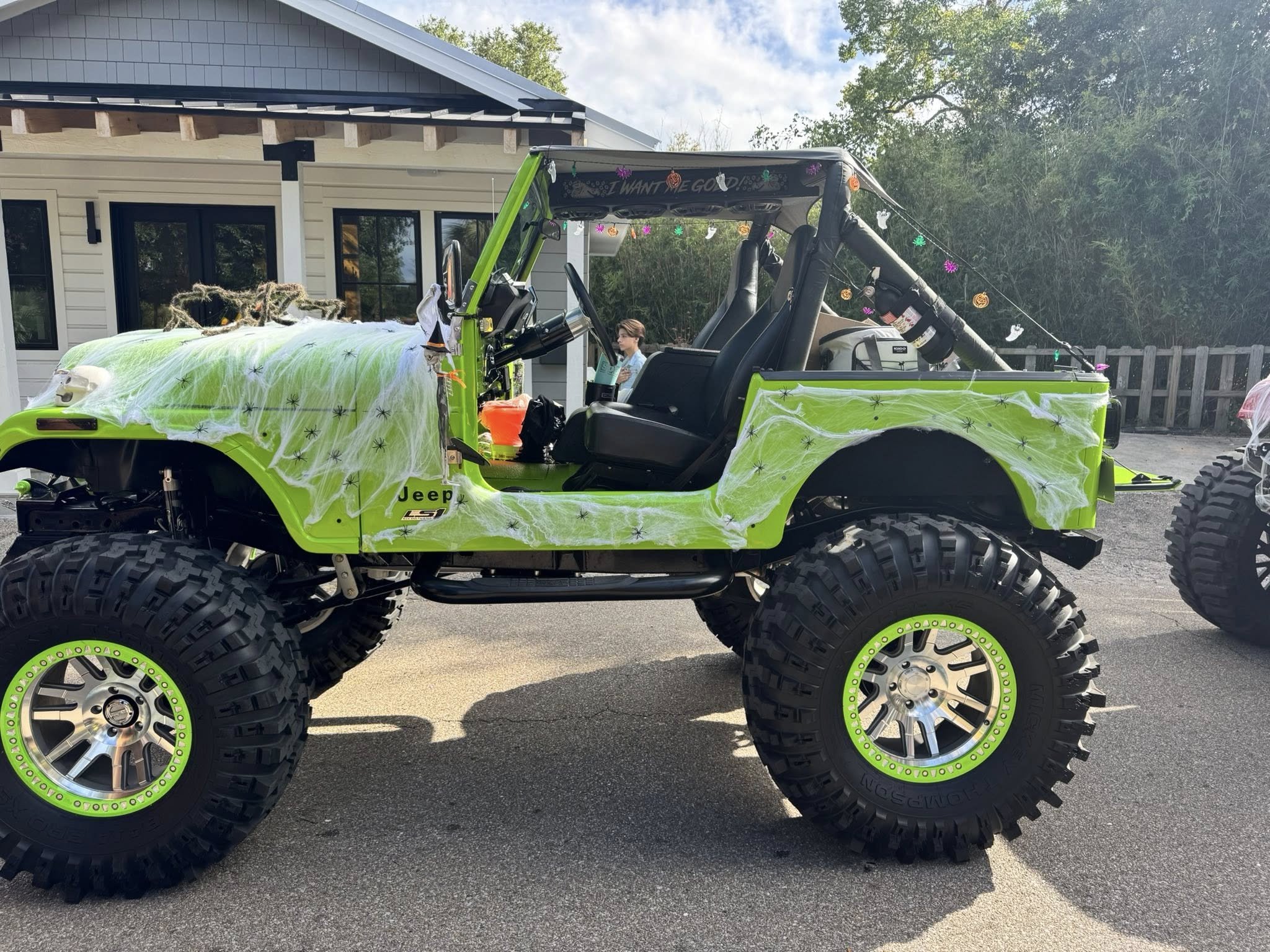 A bright green Jeep with large off-road tires decorated with Halloween cobwebs and spider decorations, parked outside a house with Halloween lights and decorations visible.