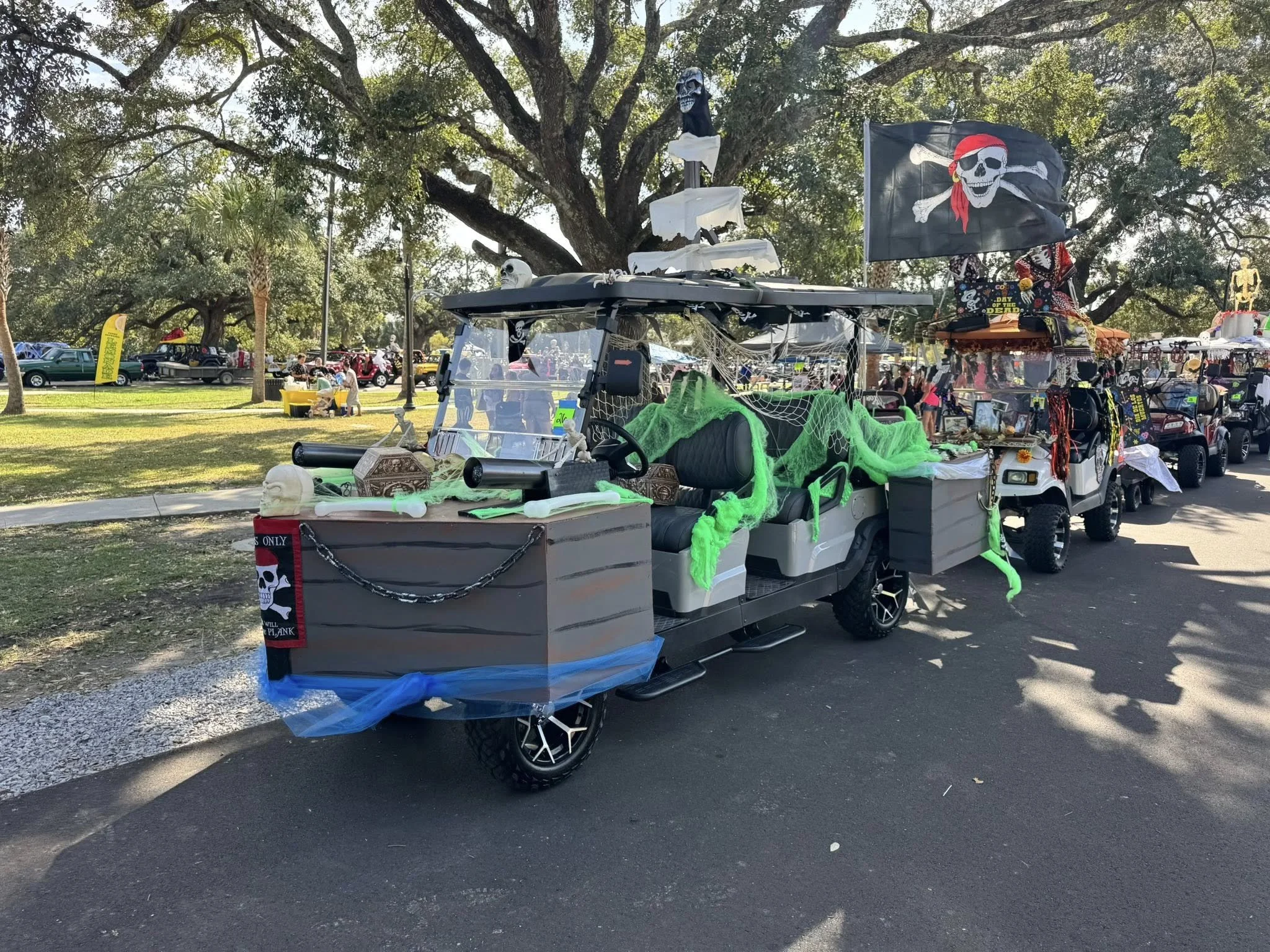 A decorated golf cart with skull and pirate-themed decorations, including a pirate flag with a skull wearing a red bandana and sunglasses, at an outdoor event with numerous cars and trees in the background.