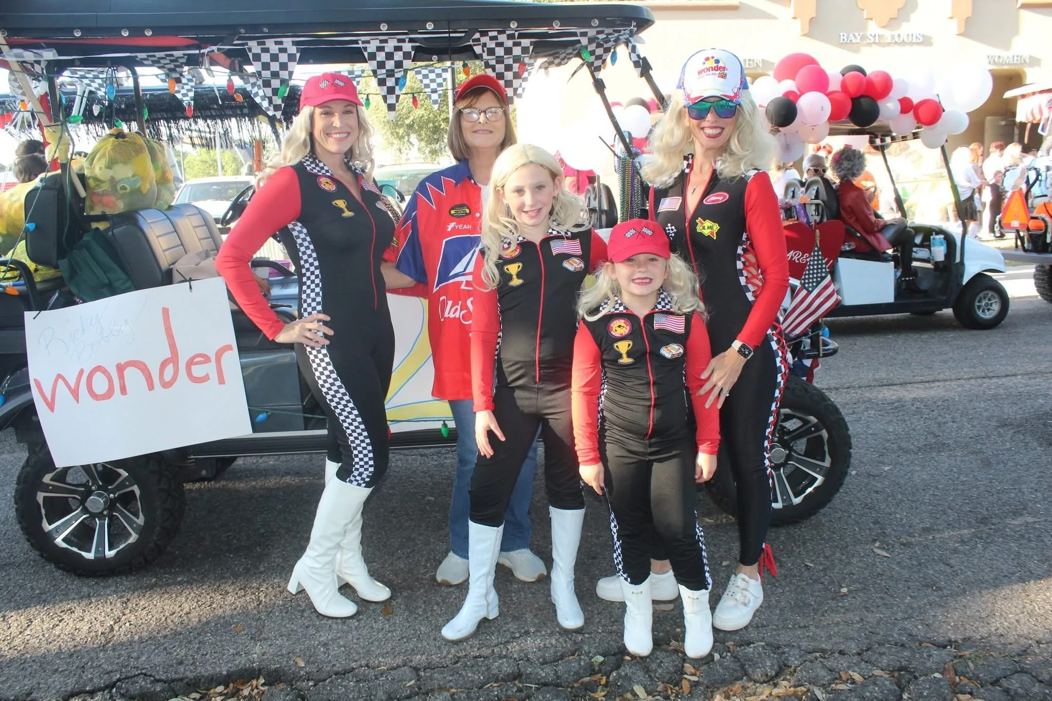 Group of five women and two young girls dressed in racing-themed outfits with checkered flags, standing in front of a decorated golf cart with balloons at a parade or festival.