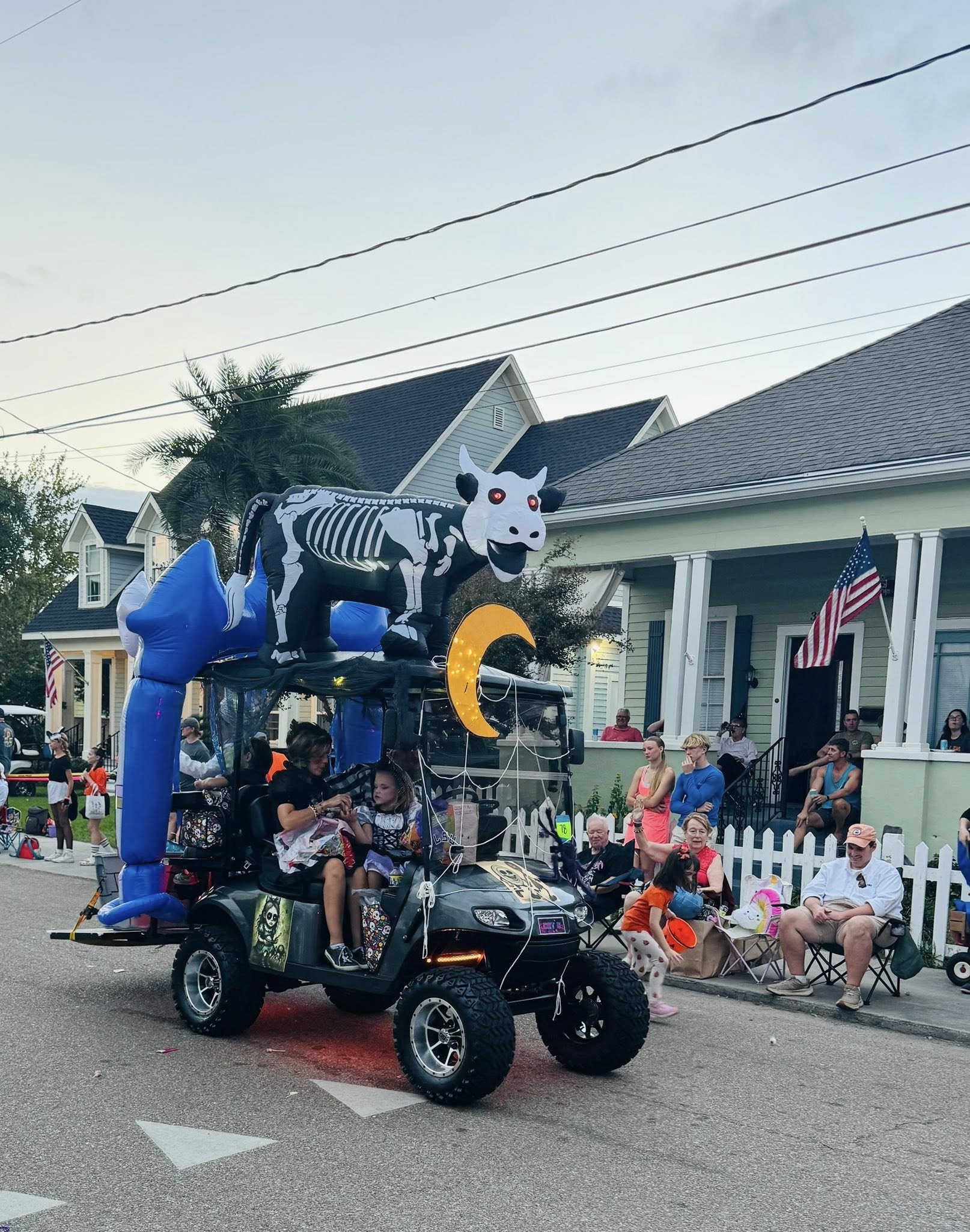 People watching a parade from the sidewalk, with a decorated golf cart featuring a cow skeleton balloon and a moon shape, and a house with American flags in the background.