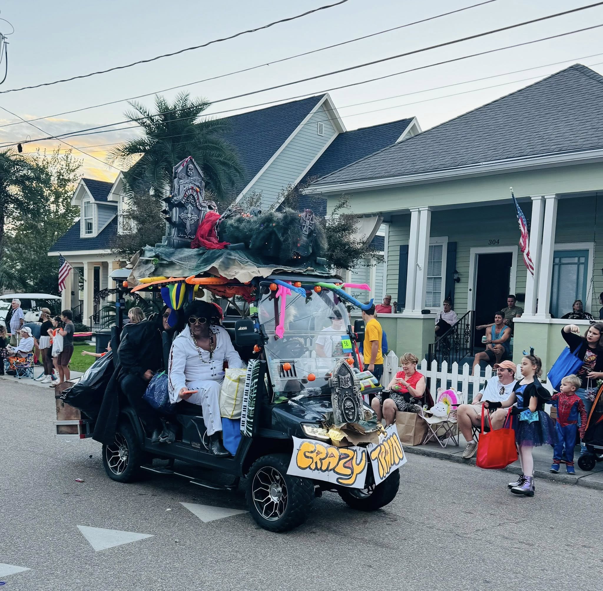 A decorated vehicle with Halloween decorations and a sign, surrounded by people on a residential street during a parade or celebration.