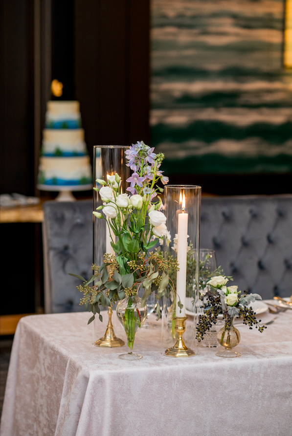 Elegant table centerpiece with white and purple flowers, candles, and gold accents on a white tablecloth in a decorated room.