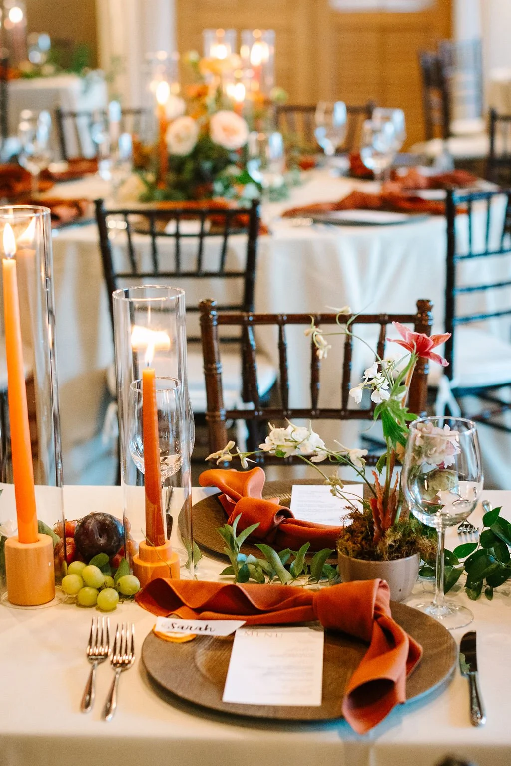 A beautifully decorated dining table set for a formal event with candles, a floral centerpiece, place settings with name cards, napkins, and wine glasses, in an elegant venue with other similarly decorated tables in the background.