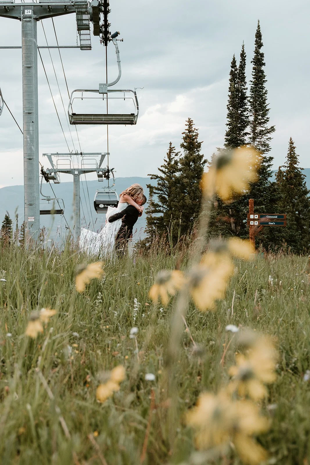 A couple hugging and kissing in a grassy field with wildflowers, ski lift chairs hanging above, and a mountain landscape with tall pine trees in the background.