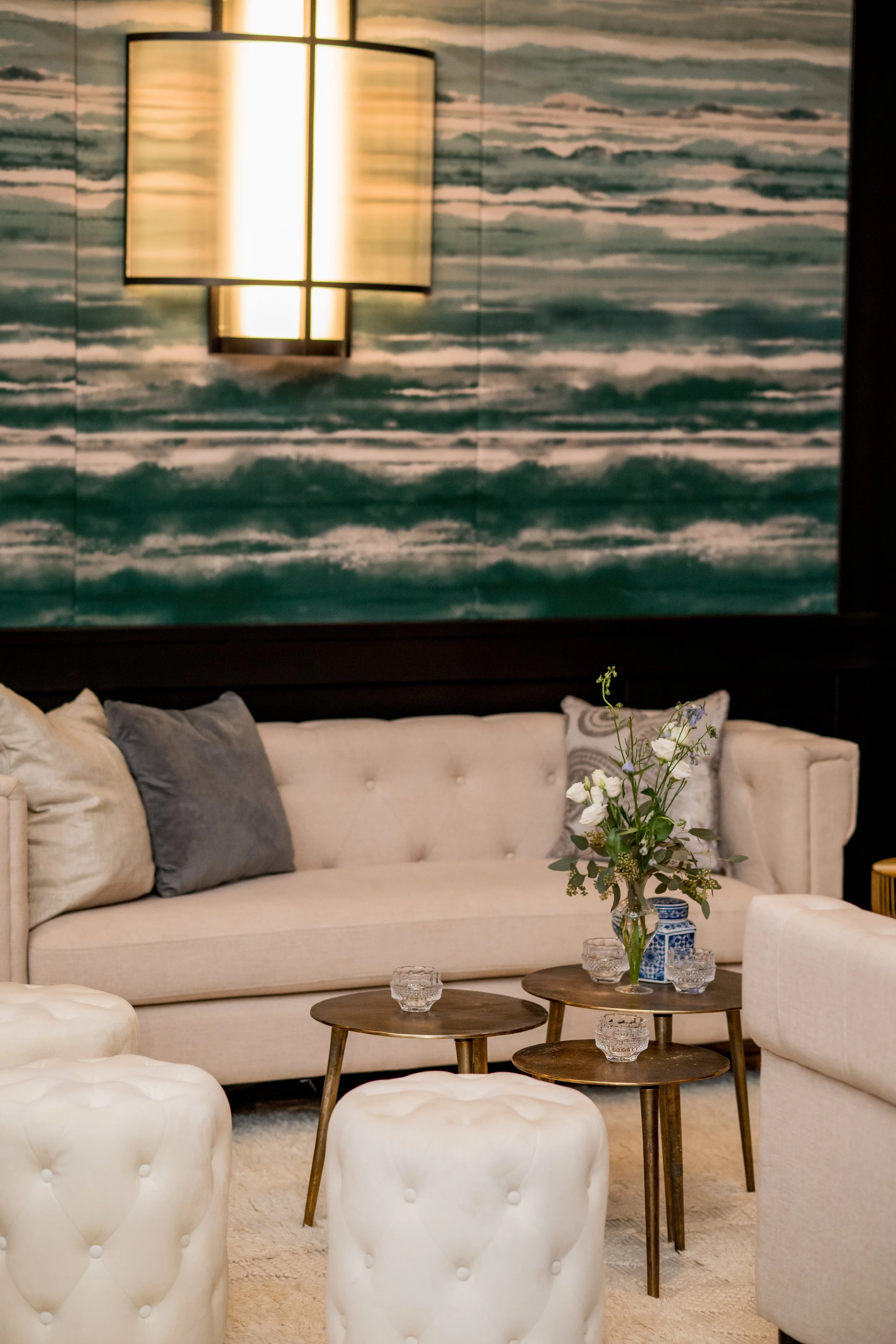 A modern living room with a beige sofa, decorative pillows, a gallery wall with wave art, a wall sconce, and a flower arrangement on small wooden tables.