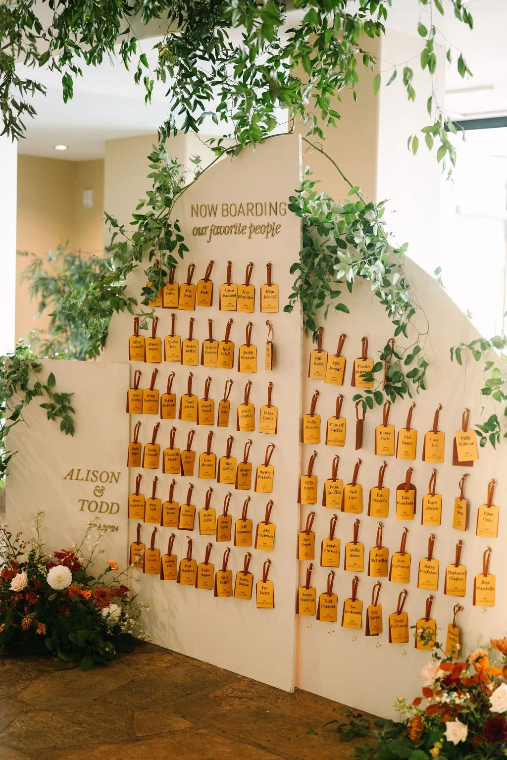A white wedding seating chart with yellow name tags hanging on leather straps, decorated with greenery and flowers, with the names Allison and Todd at the bottom.