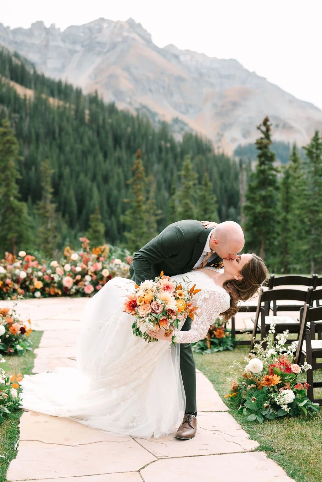 A newlywed couple sharing a kiss outdoors, with the groom leaning over the bride who is holding a large bouquet of flowers, set against a scenic mountain and forest background.