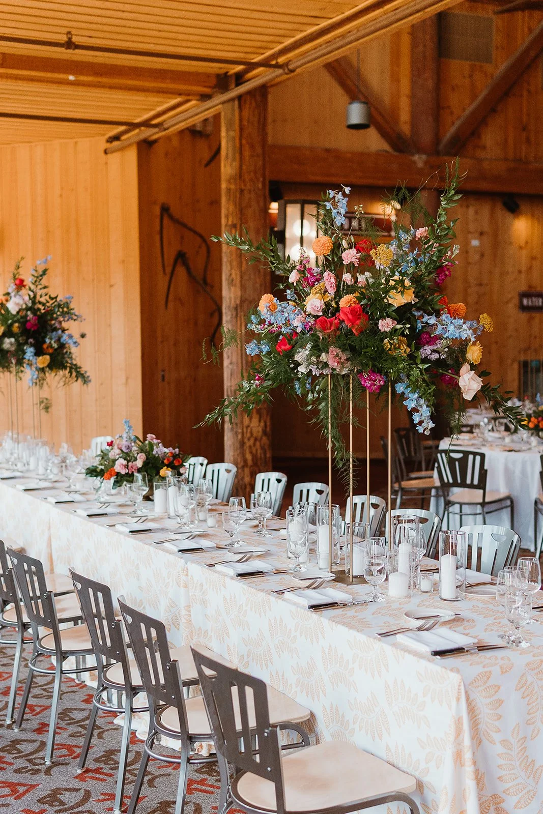 A large ski lodge table decorated with a tall, vibrant floral arrangement, set with glassware, candles, and napkins, in a rustic wooden venue.