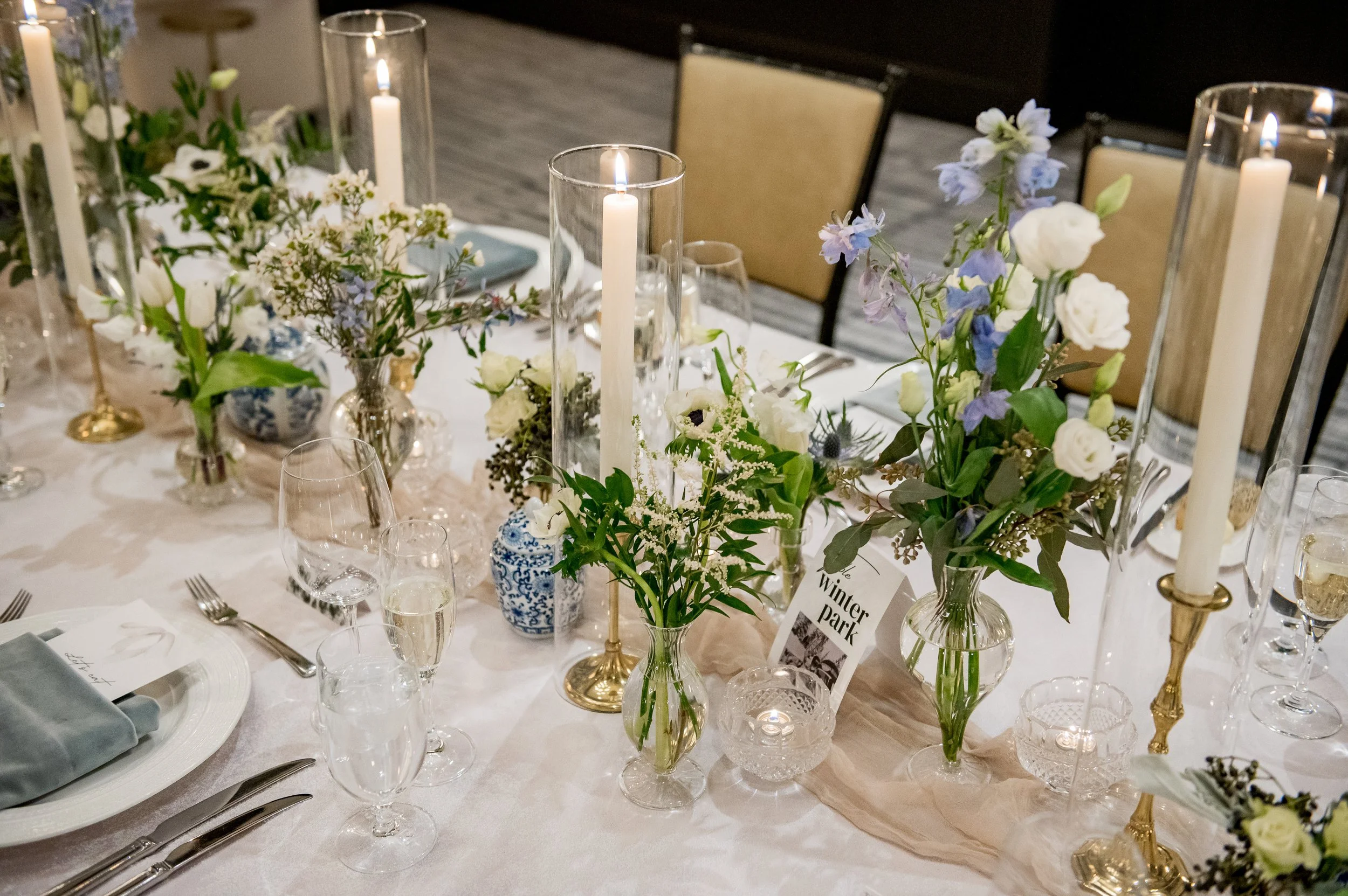 Elegant table setting with white linens, tall candle holders, and floral centerpieces including white and purple flowers.
