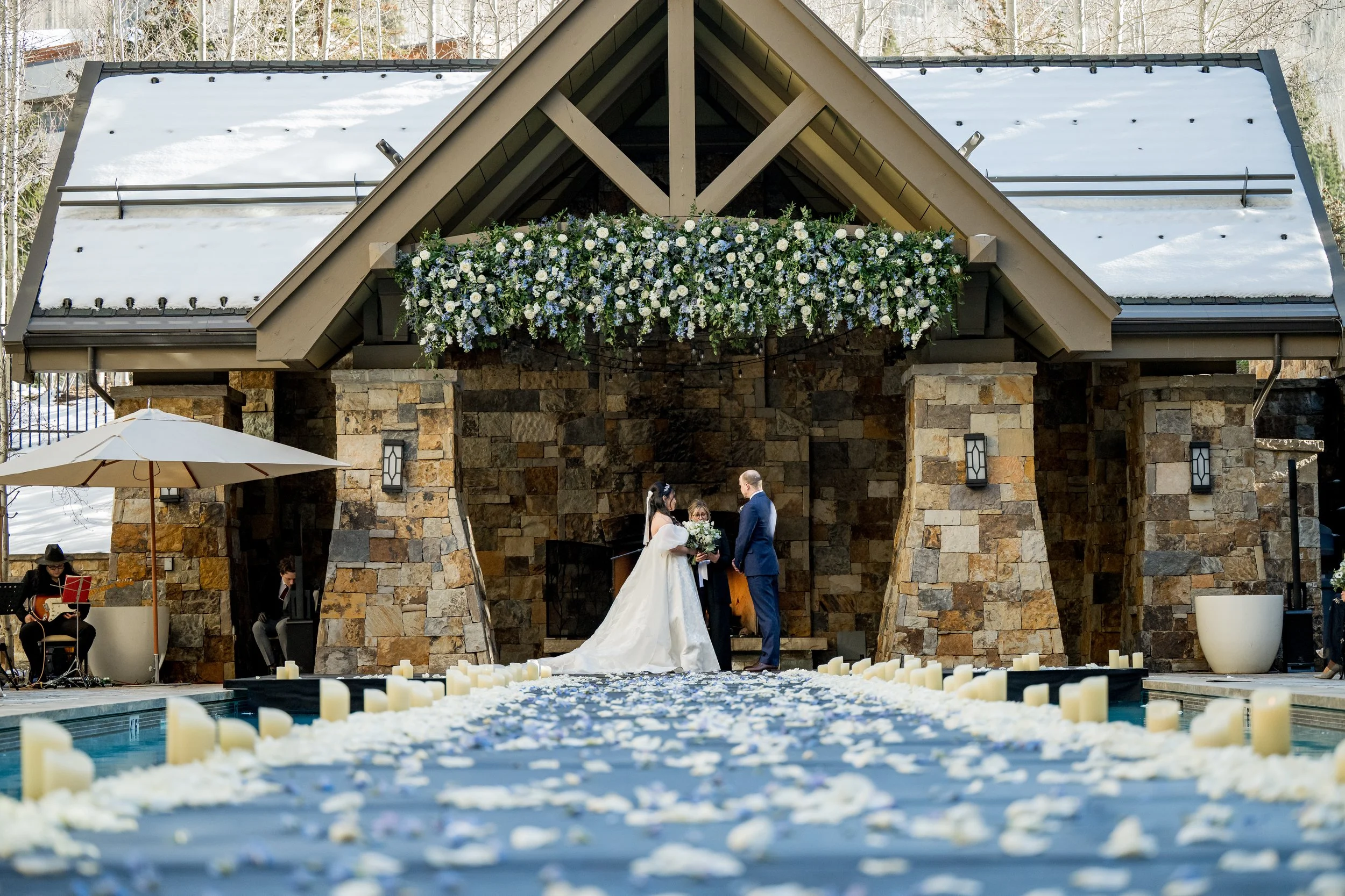 Bride and groom exchanging vows at The Four Seasons Resort & Residences Vail with a stone building backdrop, surrounded by candles and flower petals on the aisle, and musicians playing nearby.