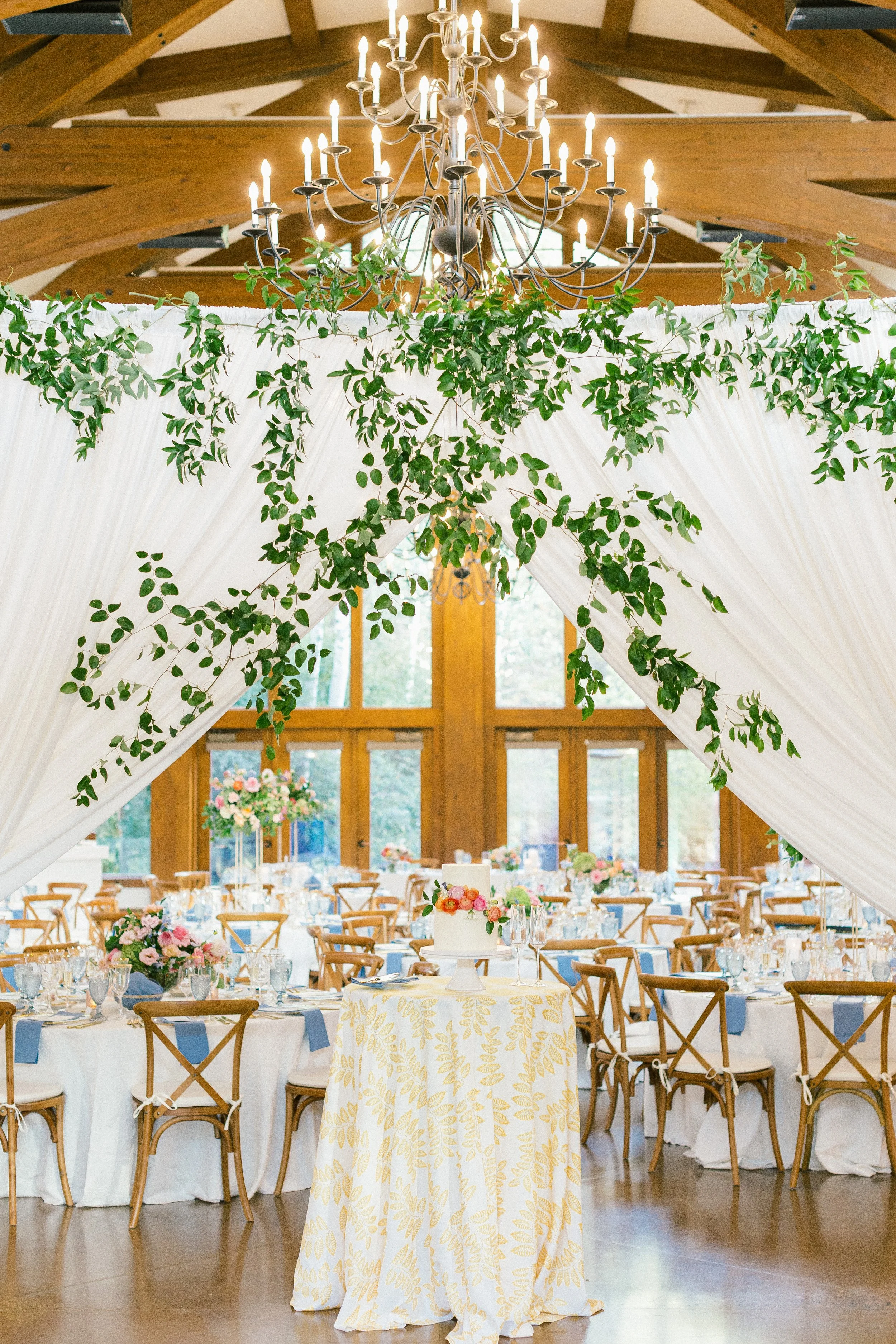 Elegant wedding reception with round tables, floral centerpieces, and a cake on a decorated table in a bright, rustic hall with large windows, green foliage, and a chandelier.
