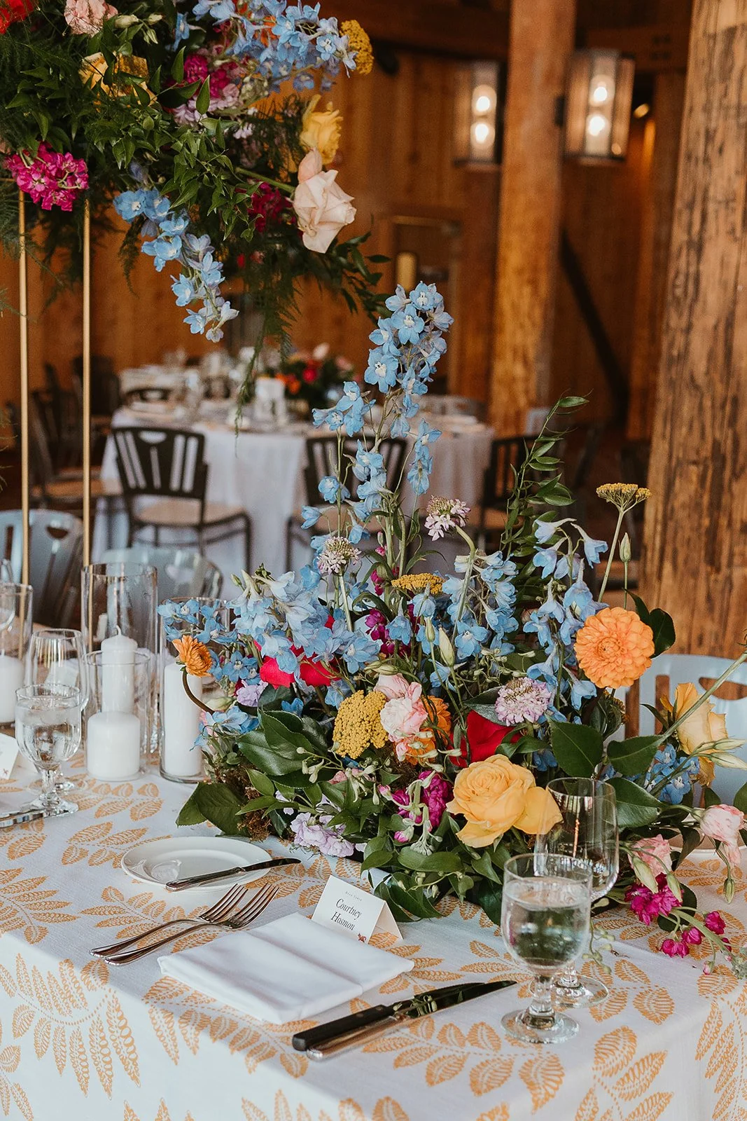 A table decorated with a large colorful floral centerpiece, white tablecloth with a gold leaf pattern, white napkins, silverware, and glassware. The background shows a rustic wooden interior with additional tables and chairs.