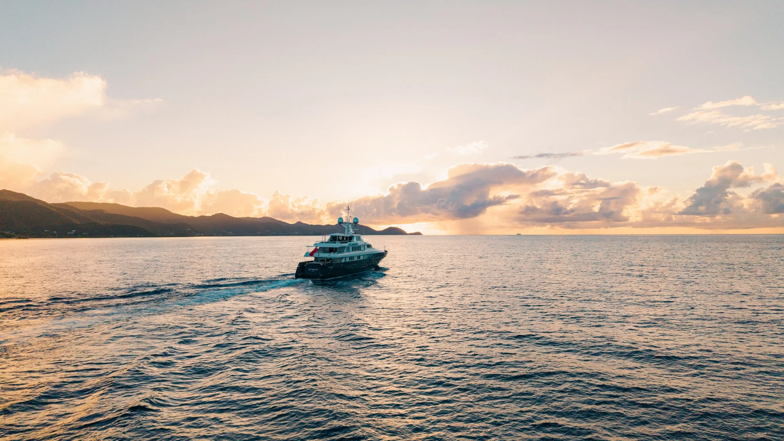 Luxury yacht on ocean during sunset with distant mountains and clouds.
