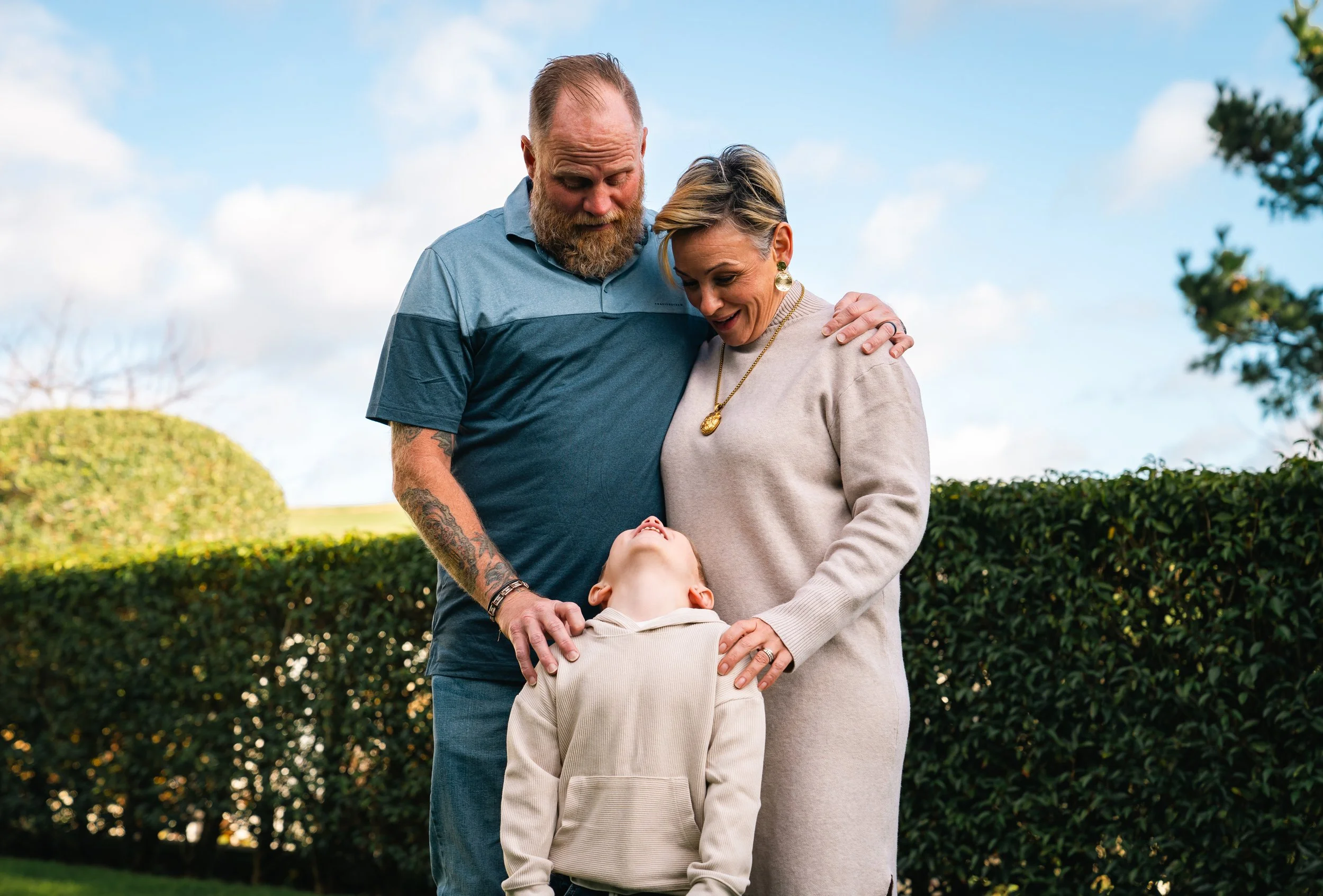 A man and woman stand outdoors with a boy between them, smiling and looking down at the child, who is looking up. They have their arms around the boy. There are bushes and a blue sky in the background.