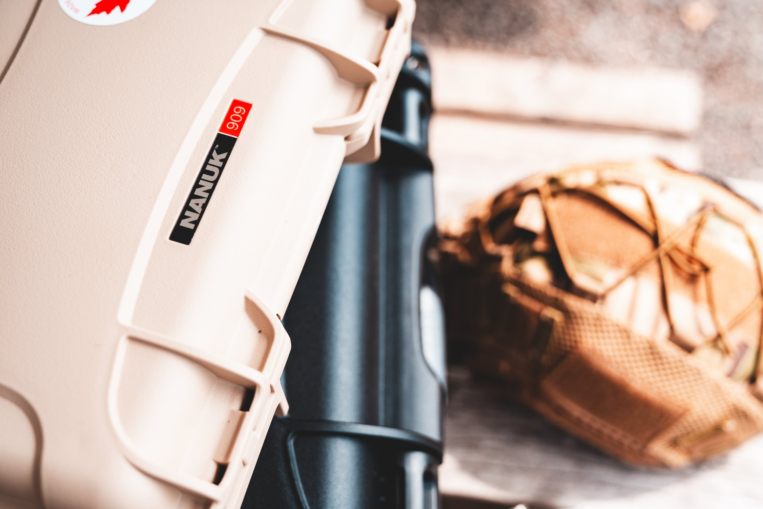 Nanuk protective case with textured beige surface, partially open, adjacent to black case and tactical backpack in blurred background.