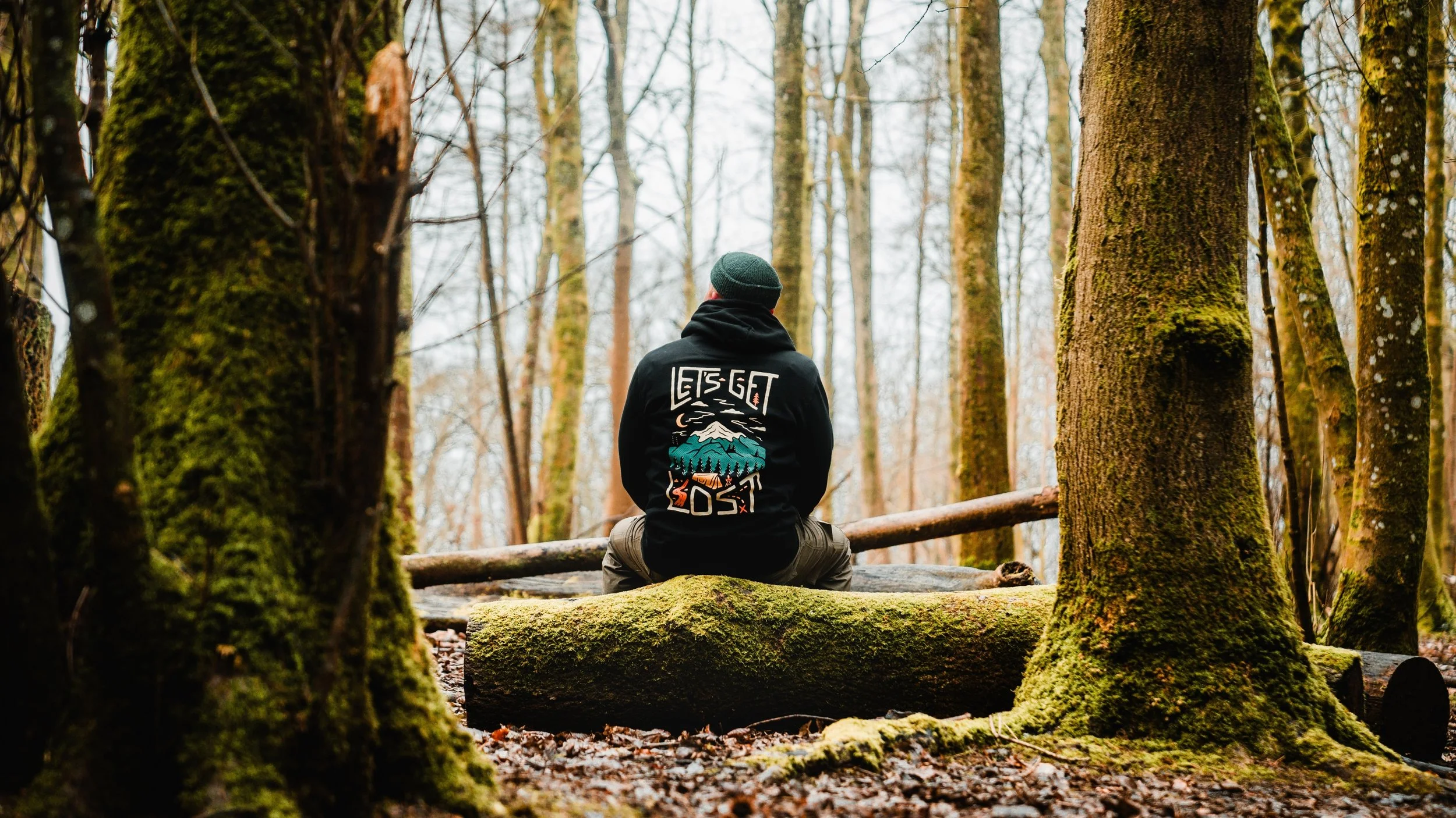 Person in a forest wearing a hoodie that reads 'Let’s Get Lost,' sitting on a mossy log.