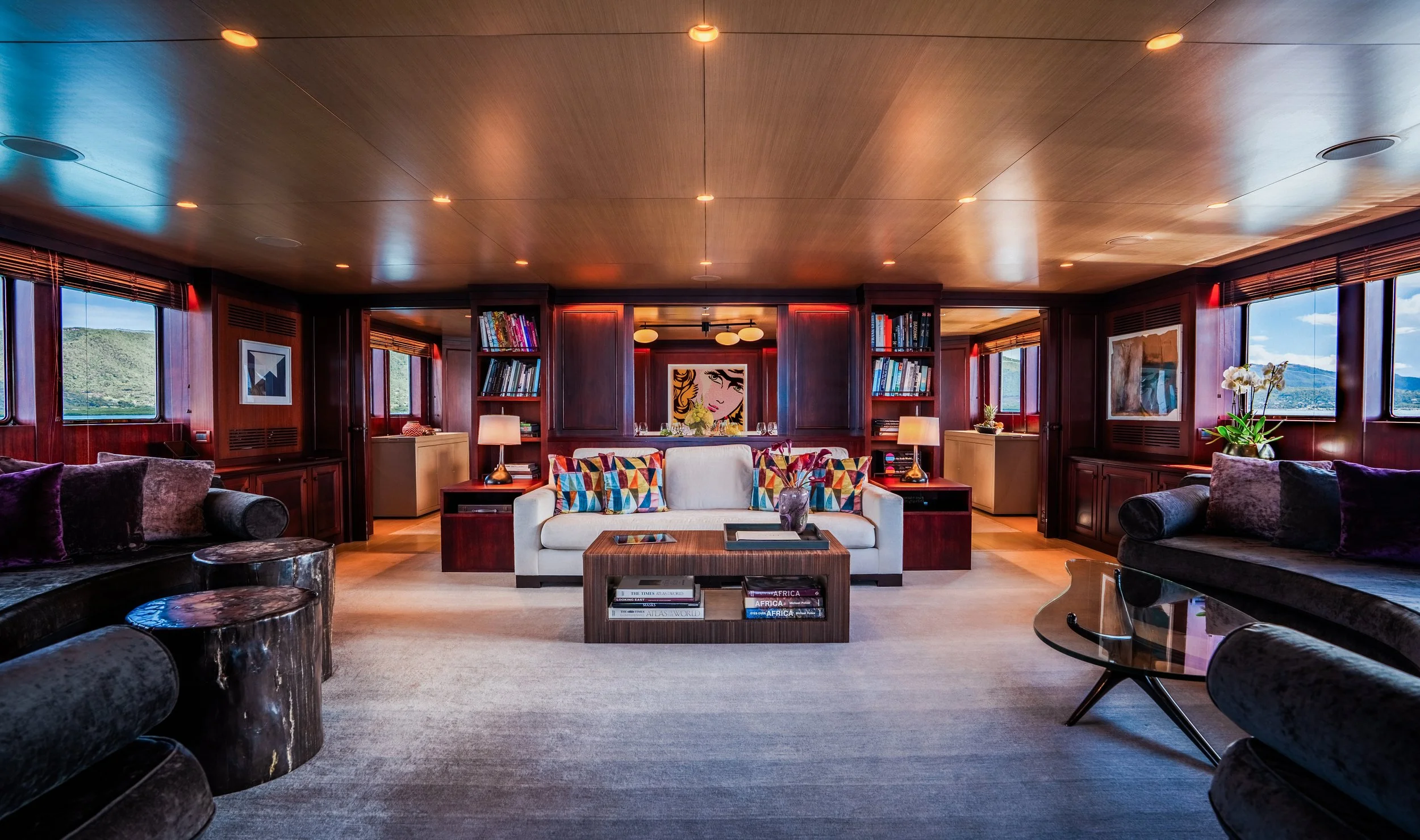 Luxurious yacht interior with wooden paneling, plush seating, colorful pillows, bookshelves, and large windows providing an ocean view.