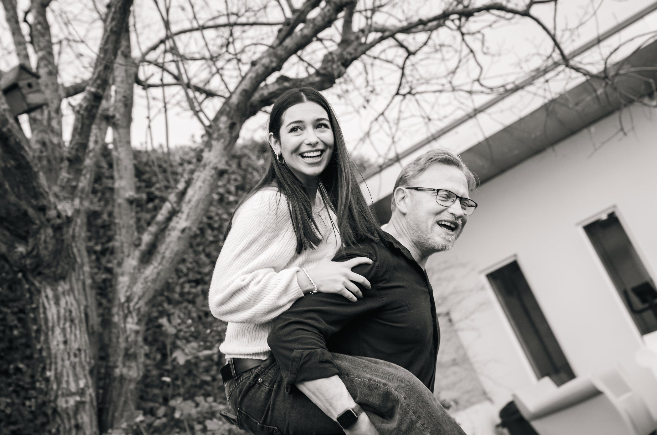 A black and white photo of a smiling woman giving a piggyback ride to a laughing man in an outdoor setting with trees and a building in the background.
