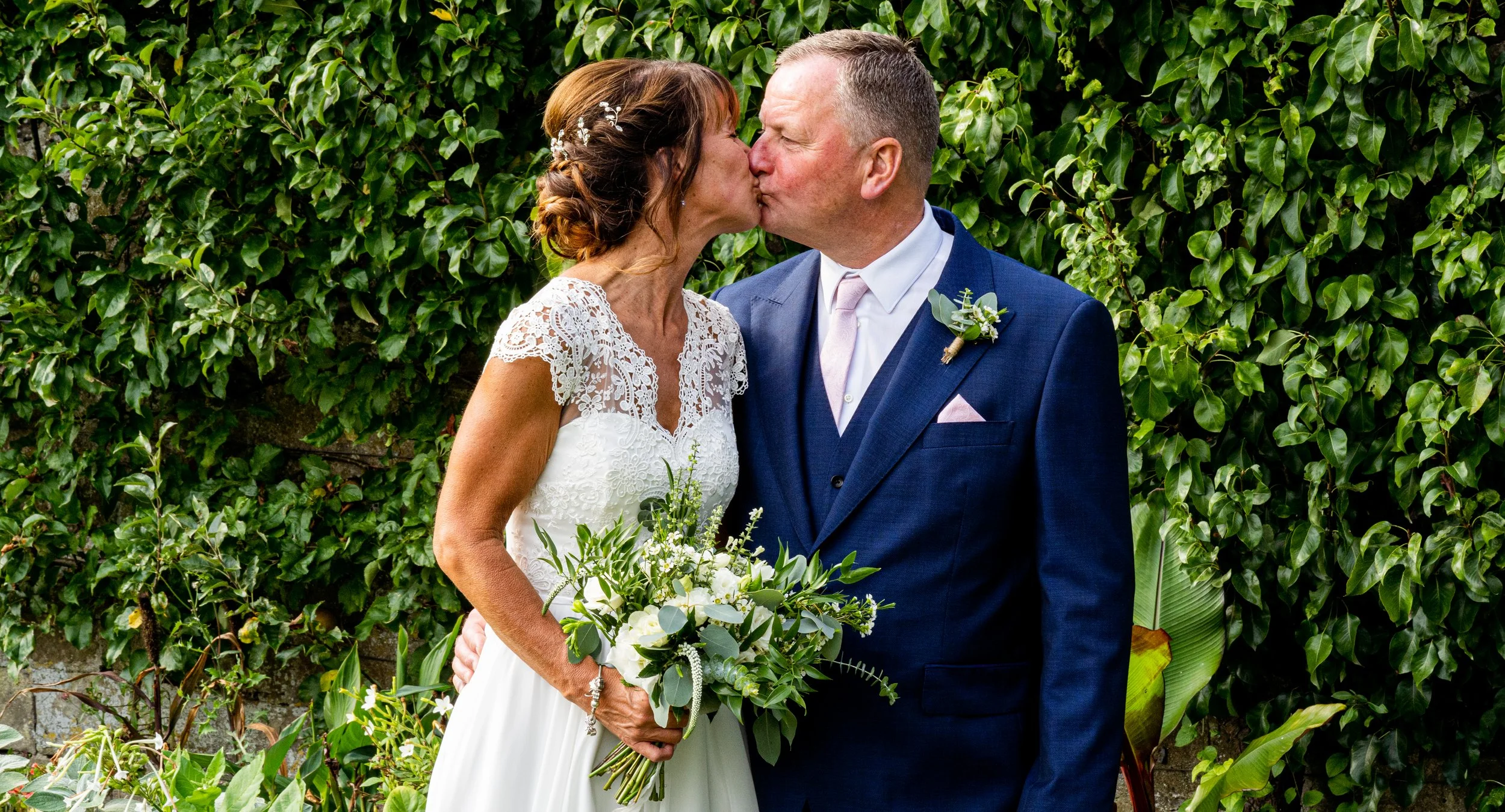 Bride and groom kissing in front of green foliage, with bride holding a bouquet of white flowers.