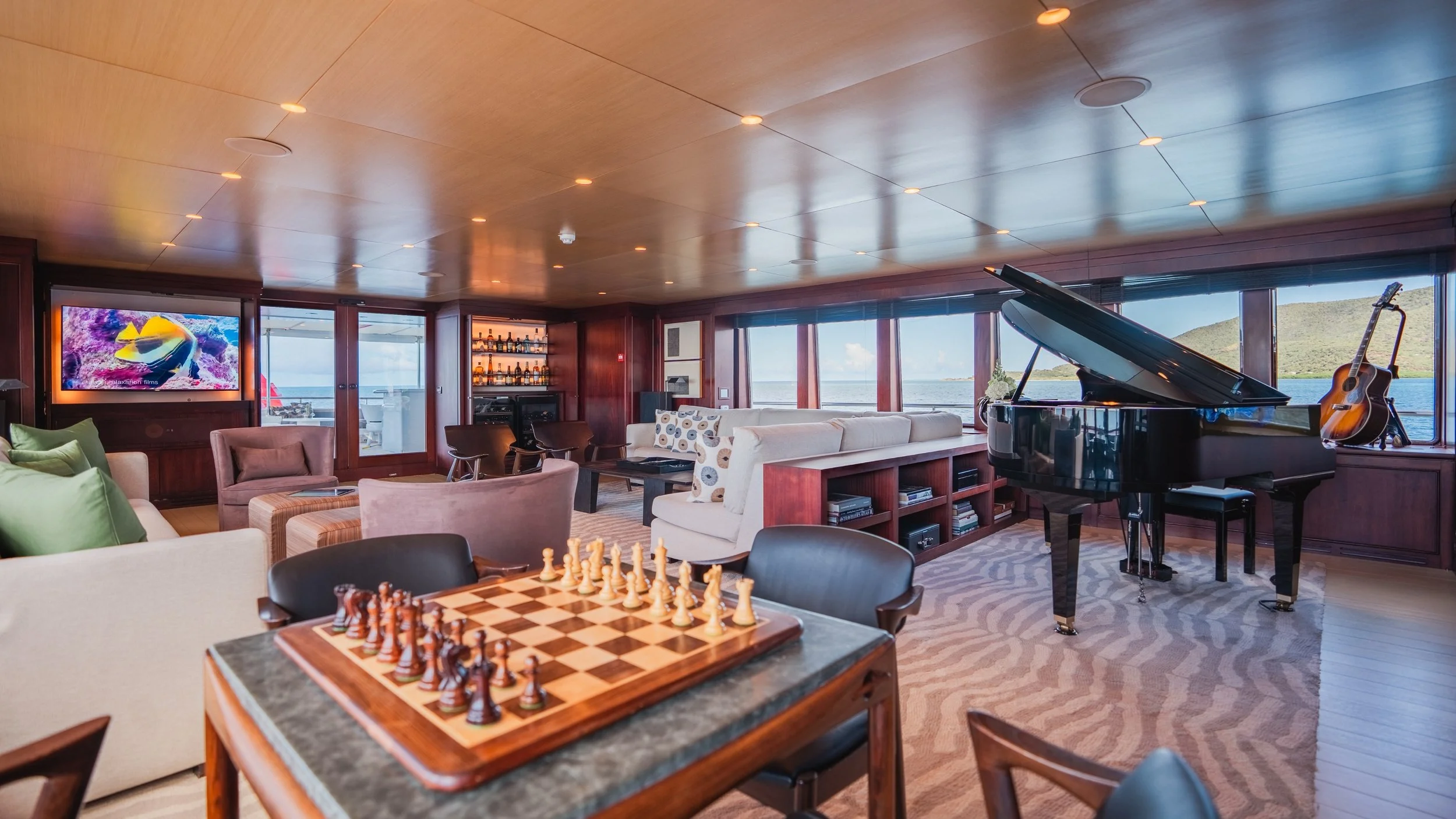 Luxury yacht interior with grand piano, chessboard, TV, and seating area overlooking ocean view.