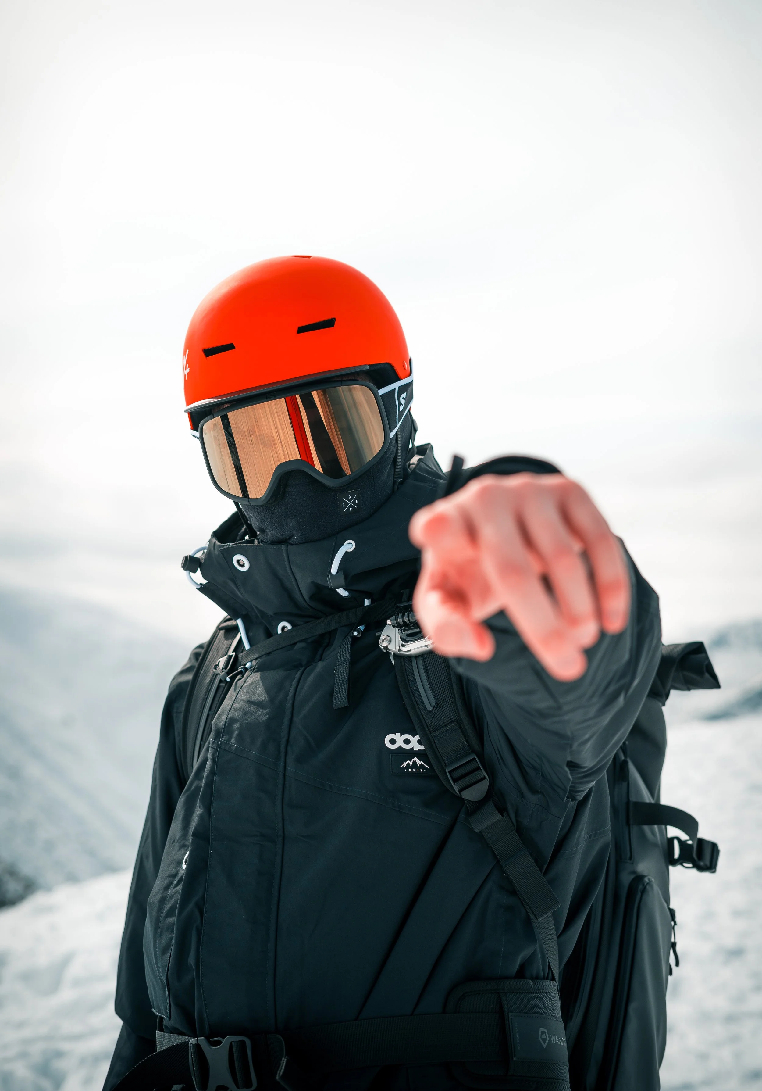 Snowboarder pointing at camera, wearing an orange helmet and black winter gear, in a snowy landscape.