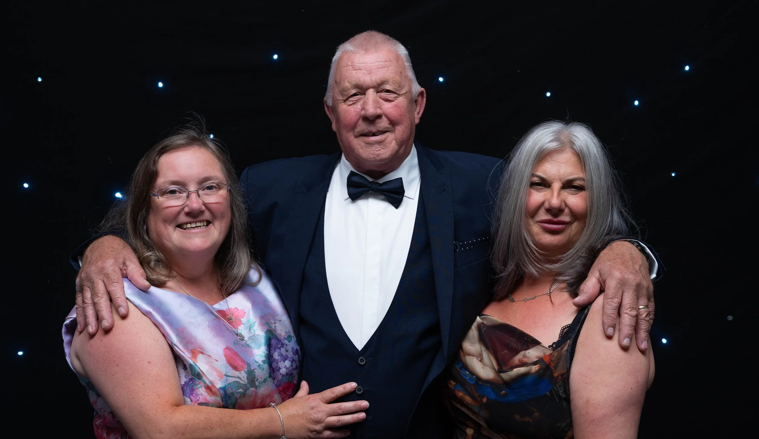 Three people posing for a formal photo, with a man in a suit and bow tie in the middle, and two women in dresses on either side. They are smiling in front of a black, starry background.