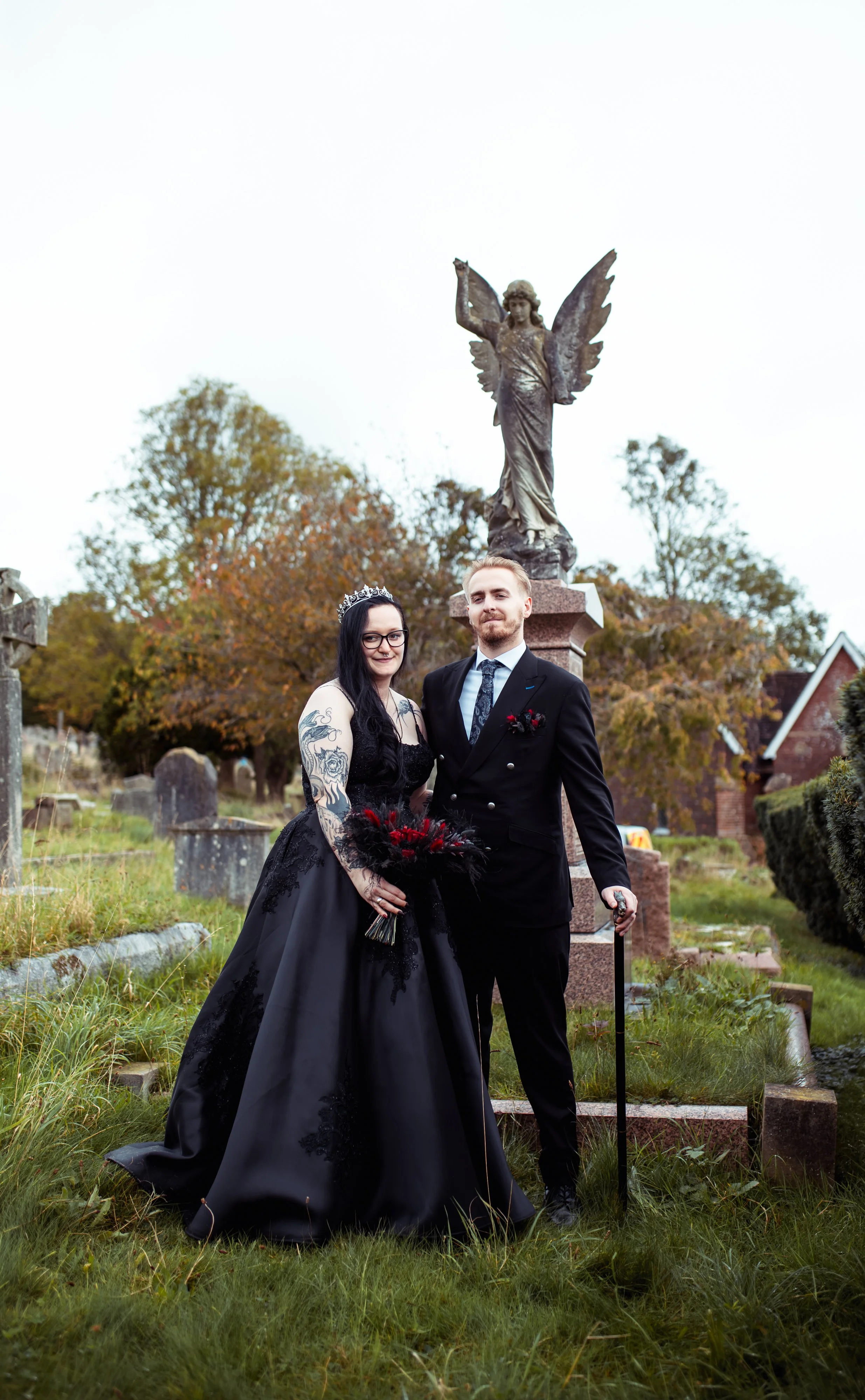 A couple in formal Gothic attire stands in a cemetery with an angel statue in the background. The woman wears a long black dress and crown, holding a bouquet, while the man is in a black suit with a cane.