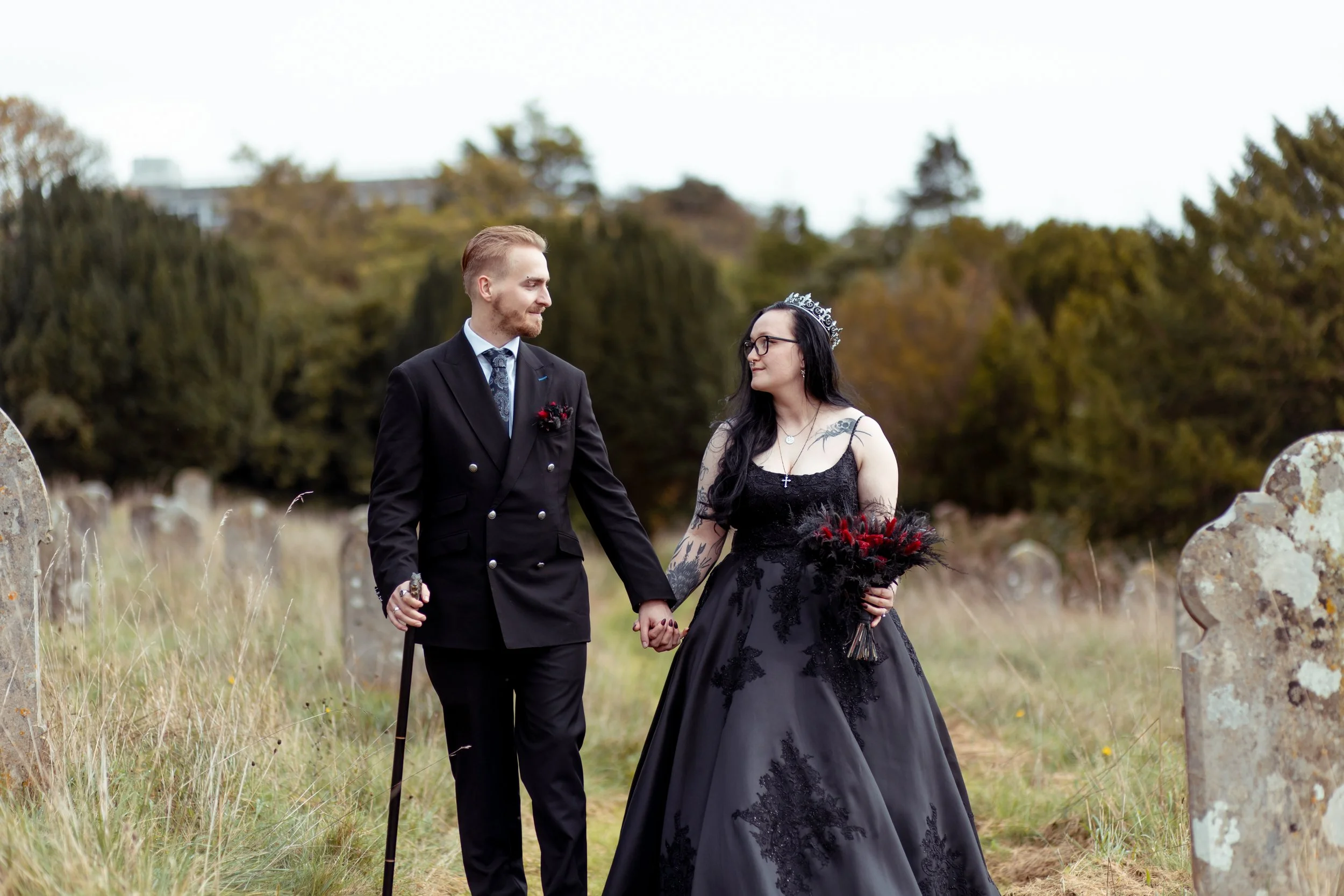 A couple in Gothic wedding attire holds hands in a cemetery, with the bride in a black dress and the groom in a black suit. They are surrounded by gravestones and trees.