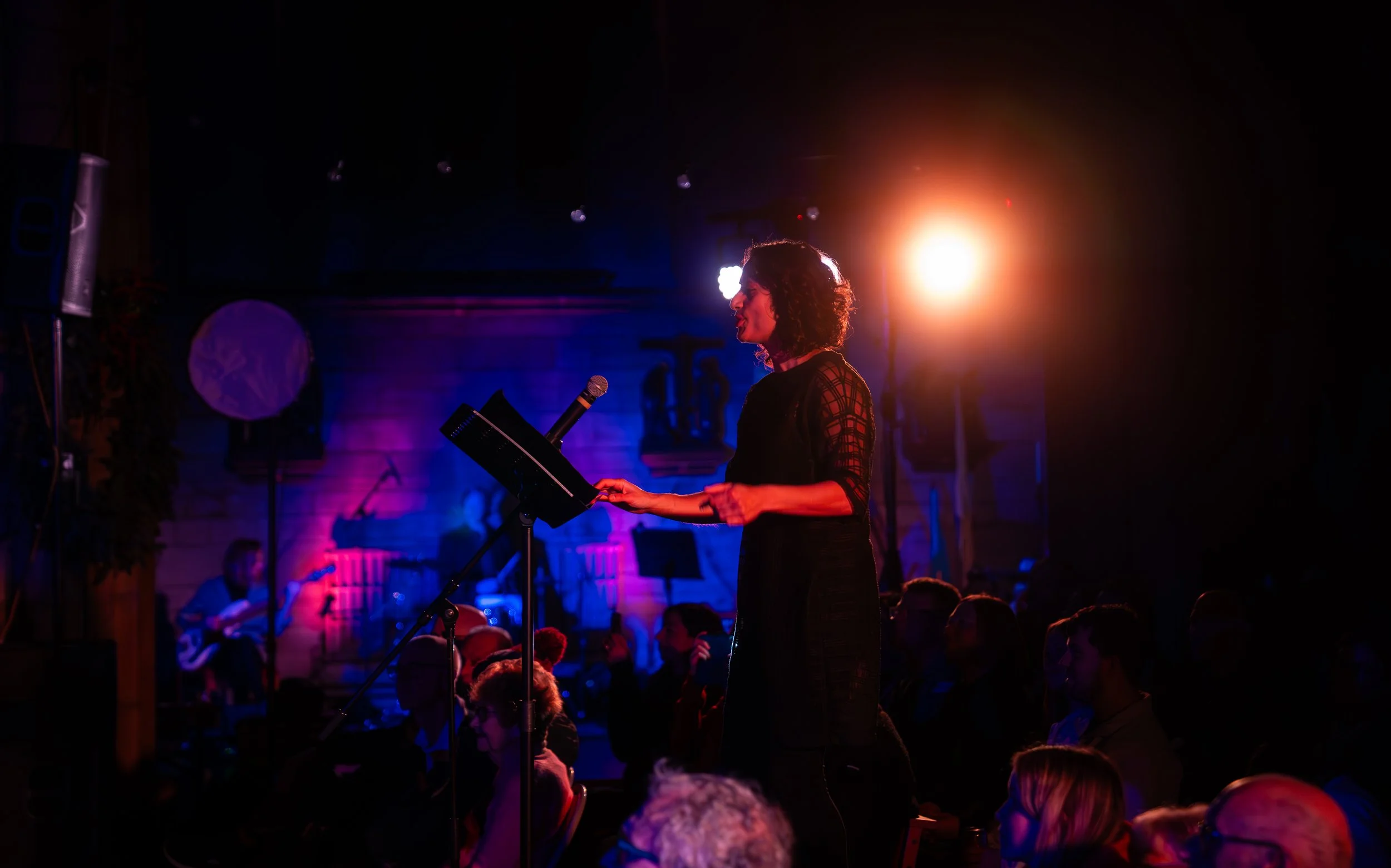 A speaker on stage with a microphone and audience in a dimly lit room with colorful lighting.