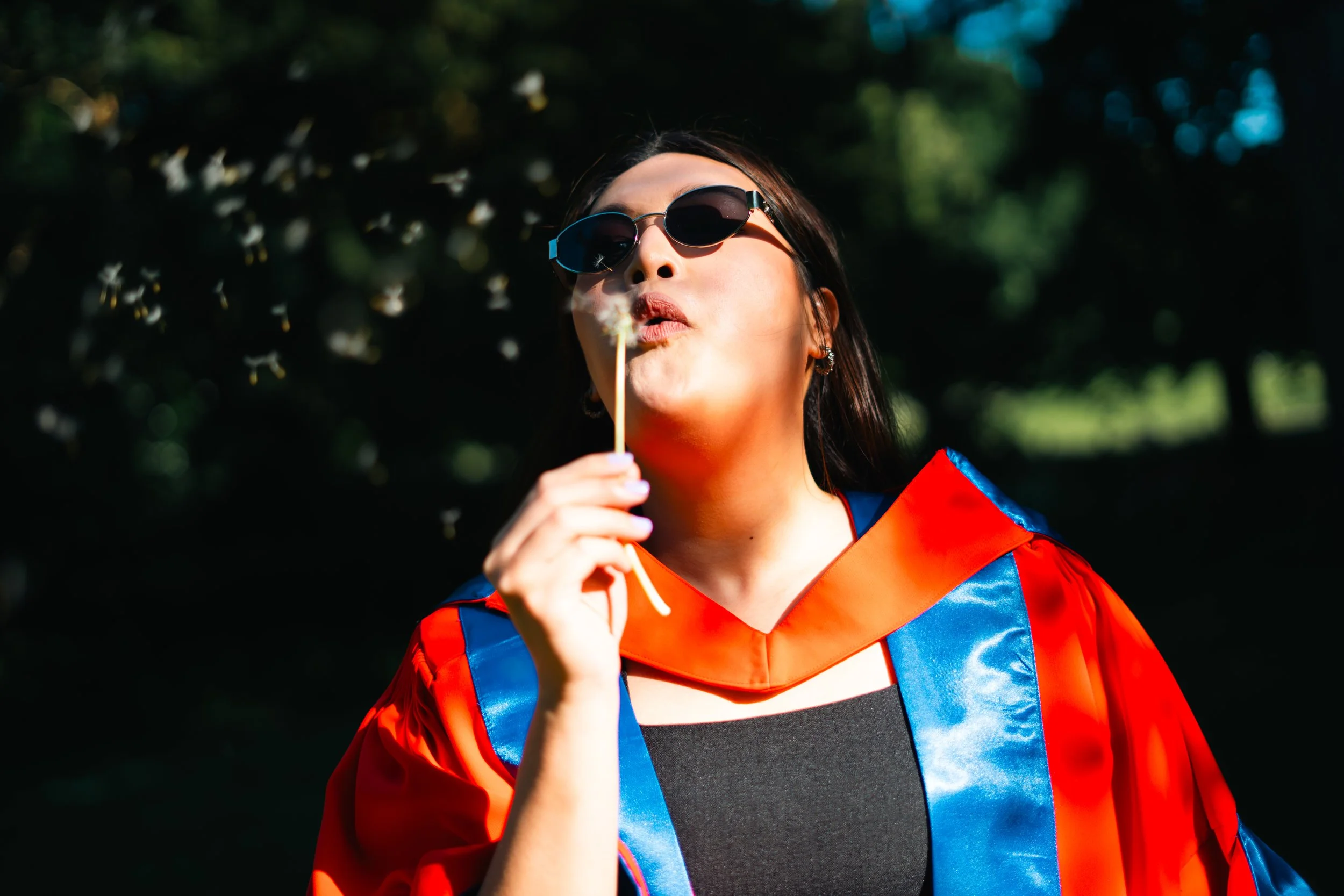 Woman in graduation gown and sunglasses holding a sparkler, outdoors with dark background and trees.