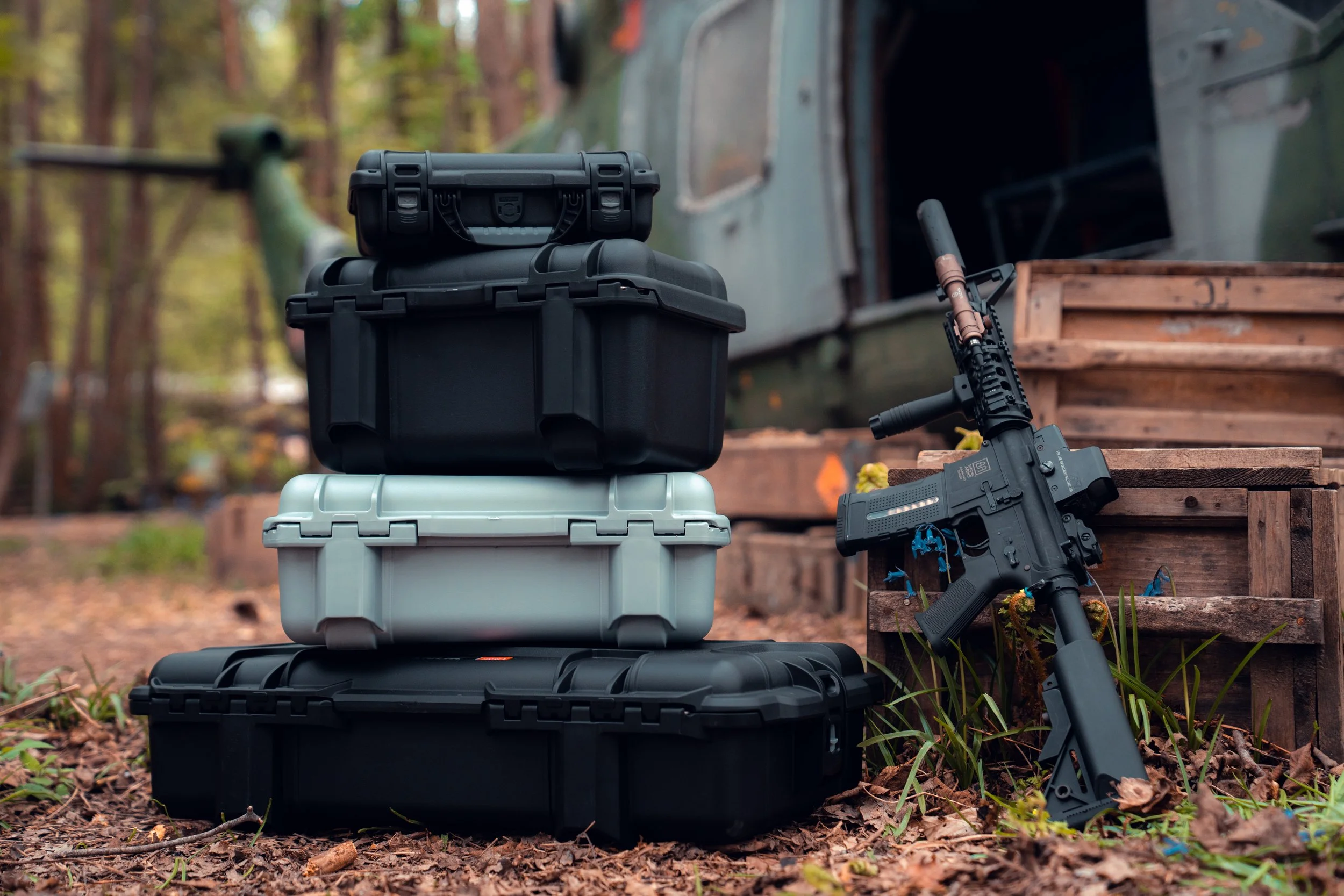 Stack of black and gray hard cases on the ground next to a rifle, with a military helicopter in the background.