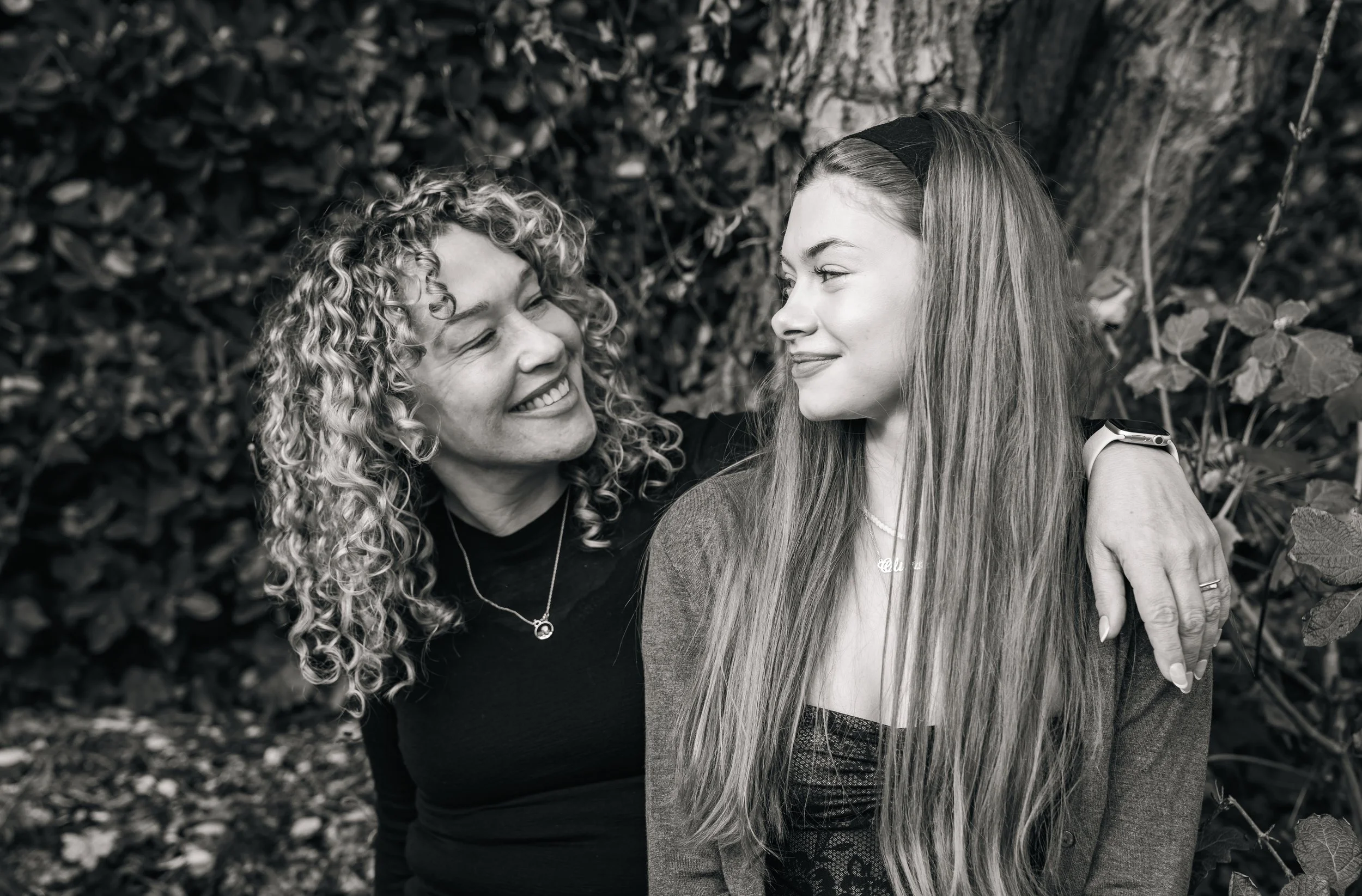 Black and white photo of two people smiling at each other, surrounded by foliage, with one person having curly hair and the other long straight hair.