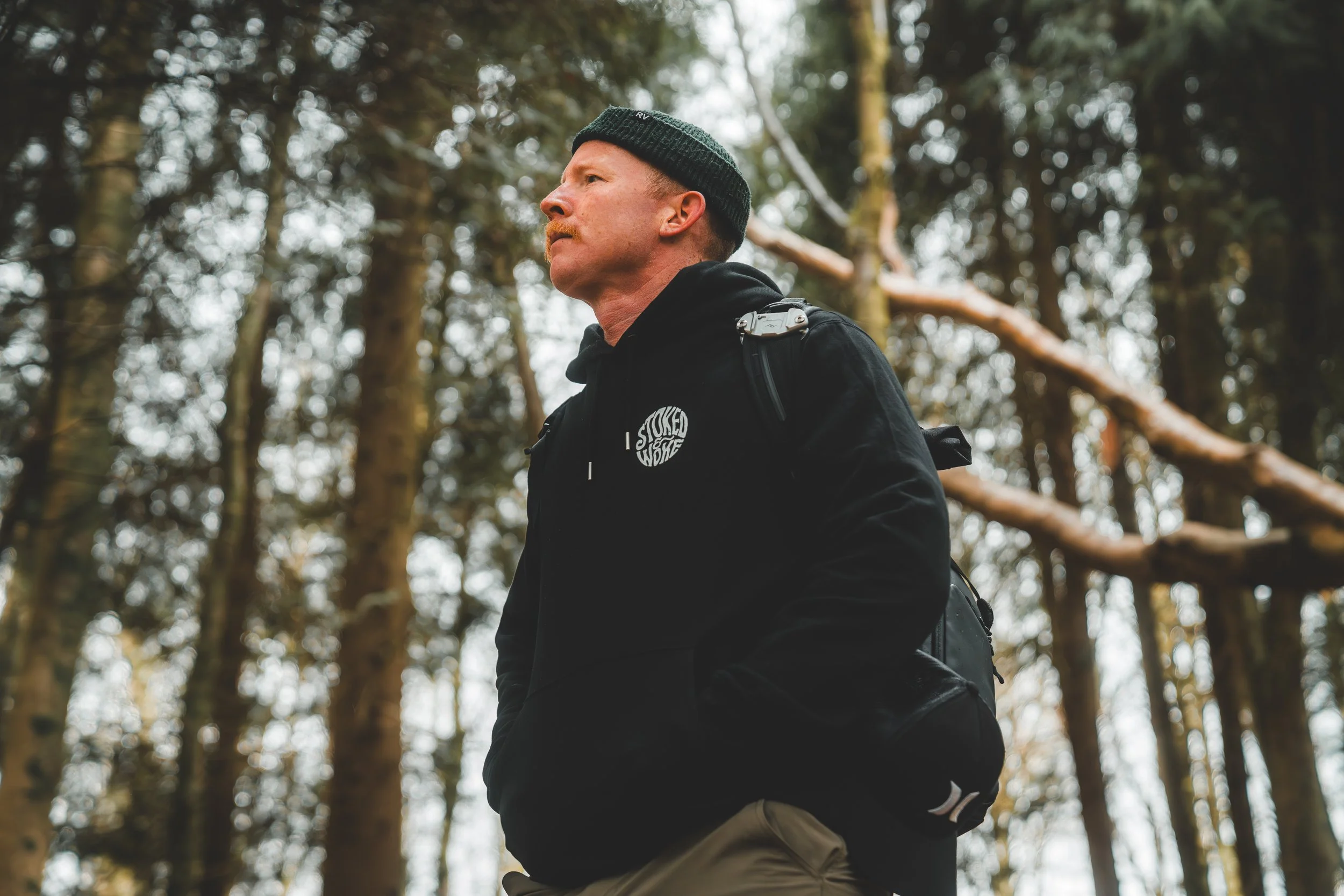 Man wearing black hoodie and hat standing in a forest with tall trees.