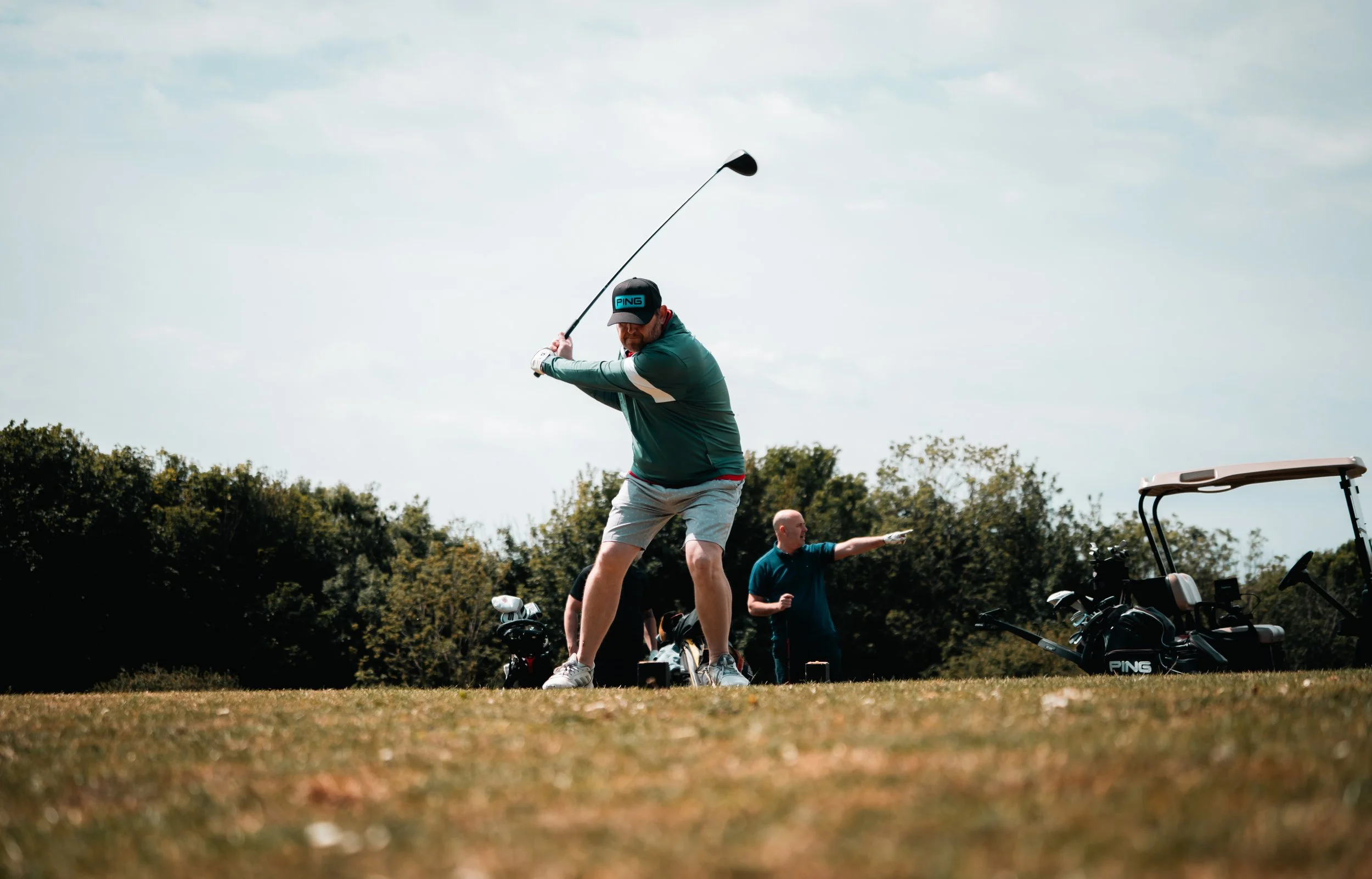 A man mid-swing playing golf on a sunny day, with two other people and golf bags in the background.
